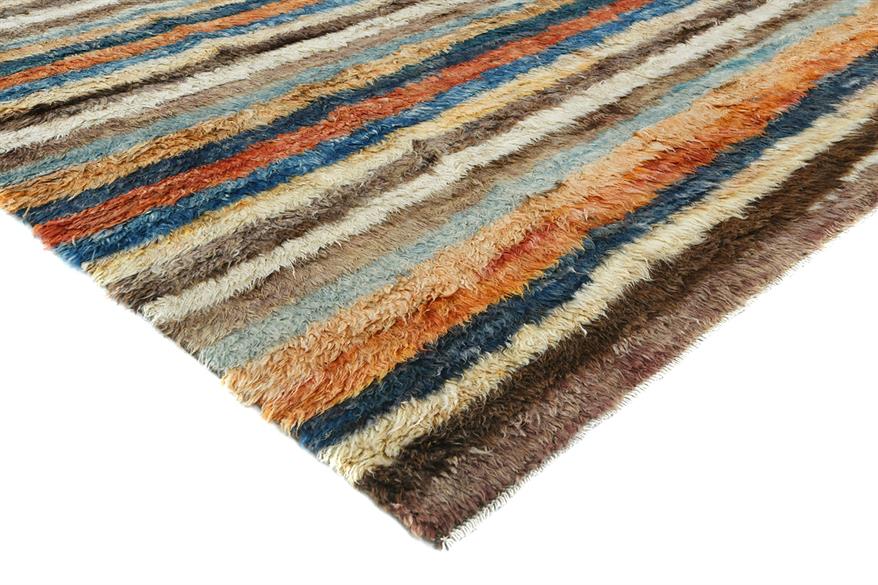 A close-up of a textured, multi-colored striped area rug with horizontal bands of blue, orange, brown, beige, and cream, shown on a plain white background. Atlantica Organics curates antique and handmade rugs, Persian, Afghan, Tibetan, Pakistani, Turkish Oushak, Kilim, Khotan, tribal and contemporary textiles for luxury interiors, homes and real estate. Our collections connect timeless craftsmanship with interior design, furniture, fine art, architecture and modern living. Explore curated rugs, collectible