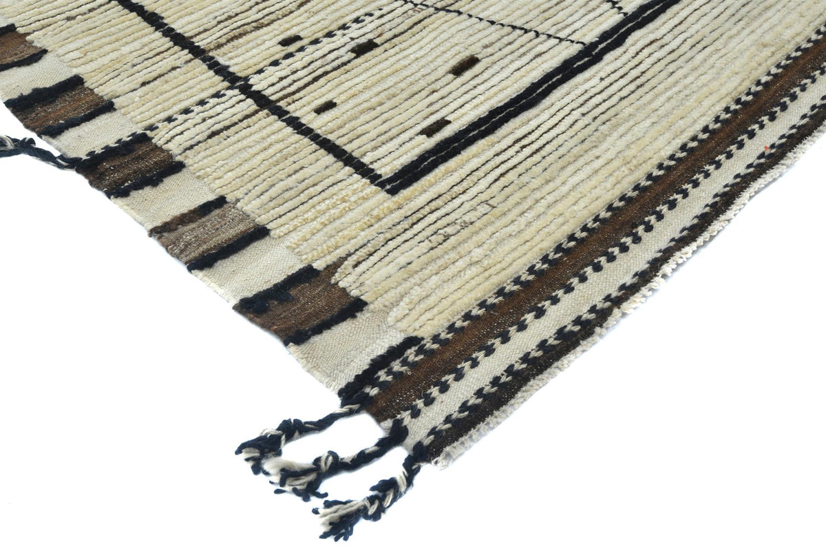 A close-up of a woven rug corner showing cream, beige, and brown stripes with black geometric patterns and fringed tassels on a white background. Atlantica Organics curates antique and handmade rugs, Persian, Afghan, Tibetan, Pakistani, Turkish Oushak, Kilim, Khotan, tribal and contemporary textiles for luxury interiors, homes and real estate. Our collections connect timeless craftsmanship with interior design, furniture, fine art, architecture and modern living. Explore curated rugs, collectible textiles,