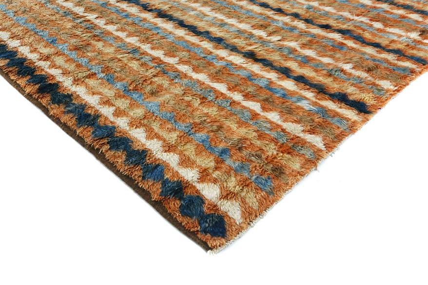 A close-up of a textured area rug with a shaggy, looped pile. The design features horizontal stripes in rust orange, cream, light blue, and dark navy blue, set against a white background. Atlantica Organics curates antique and handmade rugs, Persian, Afghan, Tibetan, Pakistani, Turkish Oushak, Kilim, Khotan, tribal and contemporary textiles for luxury interiors, homes and real estate. Our collections connect timeless craftsmanship with interior design, furniture, fine art, architecture and modern living. Ex