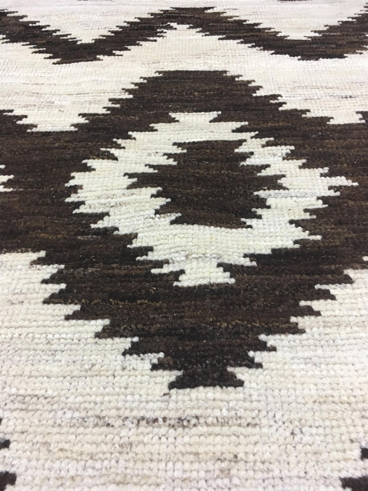 A close-up of a woven rug with a geometric diamond pattern in dark brown and off-white. Atlantica Organics curates antique and handmade rugs, Persian, Afghan, Tibetan, Pakistani, Turkish Oushak, Kilim, Khotan, tribal and contemporary textiles for luxury interiors, homes and real estate. Our collections connect timeless craftsmanship with interior design, furniture, fine art, architecture and modern living. Explore curated rugs, collectible textiles, natural materials, cultural heritage, sustainable design a