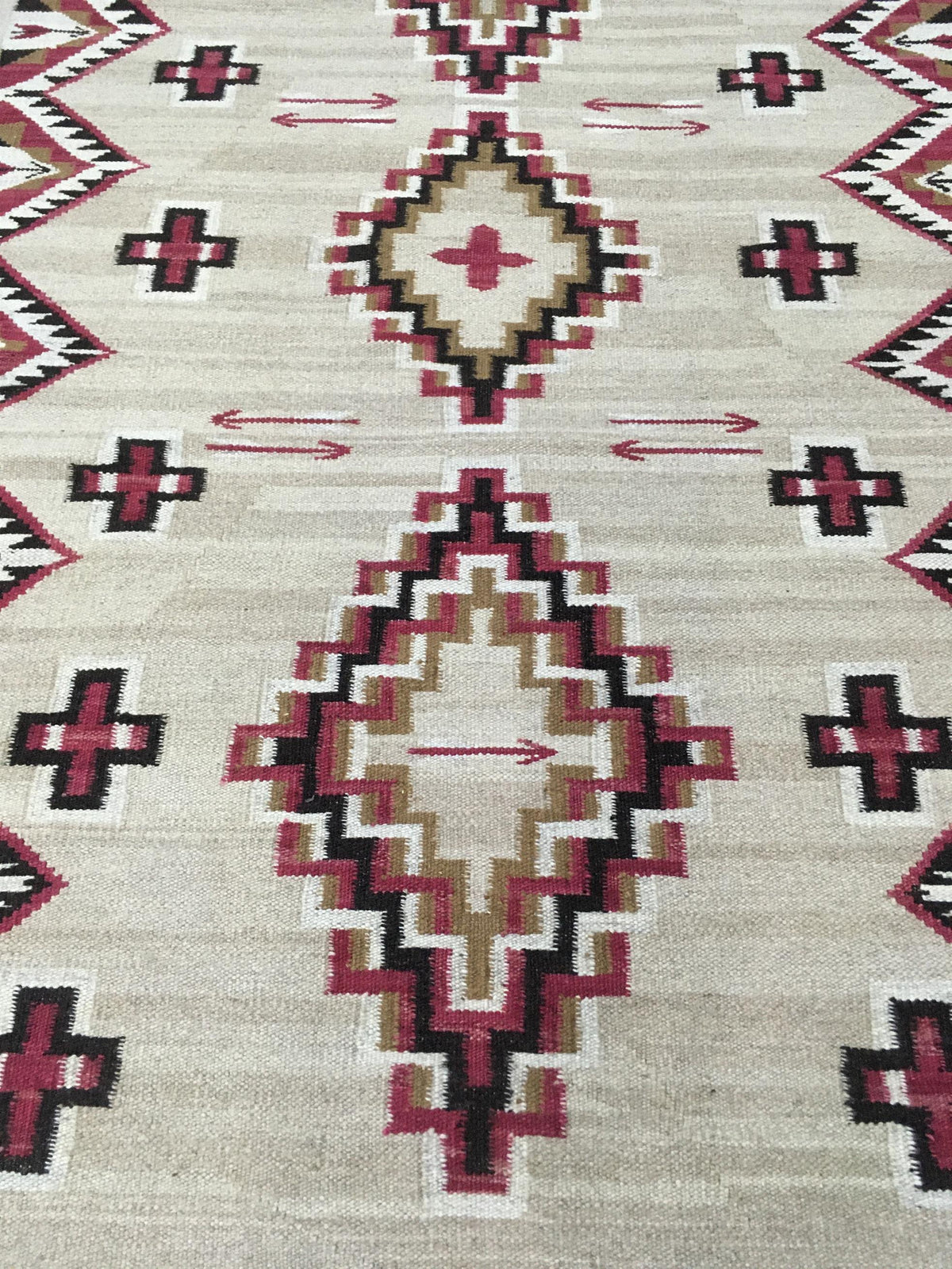 A close-up of a woven rug with a Southwestern geometric pattern. The rug features a warm beige background with large central diamond medallions and smaller diamond motifs in dark red, black, and tan, accented by small red arrows.Atlantica Organics curates antique and handmade rugs, Persian, Afghan, Tibetan, Pakistani, Turkish Oushak, Kilim, Khotan, tribal and contemporary textiles for luxury interiors, homes and real estate. Our collections connect timeless craftsmanship with interior design, furniture, fin