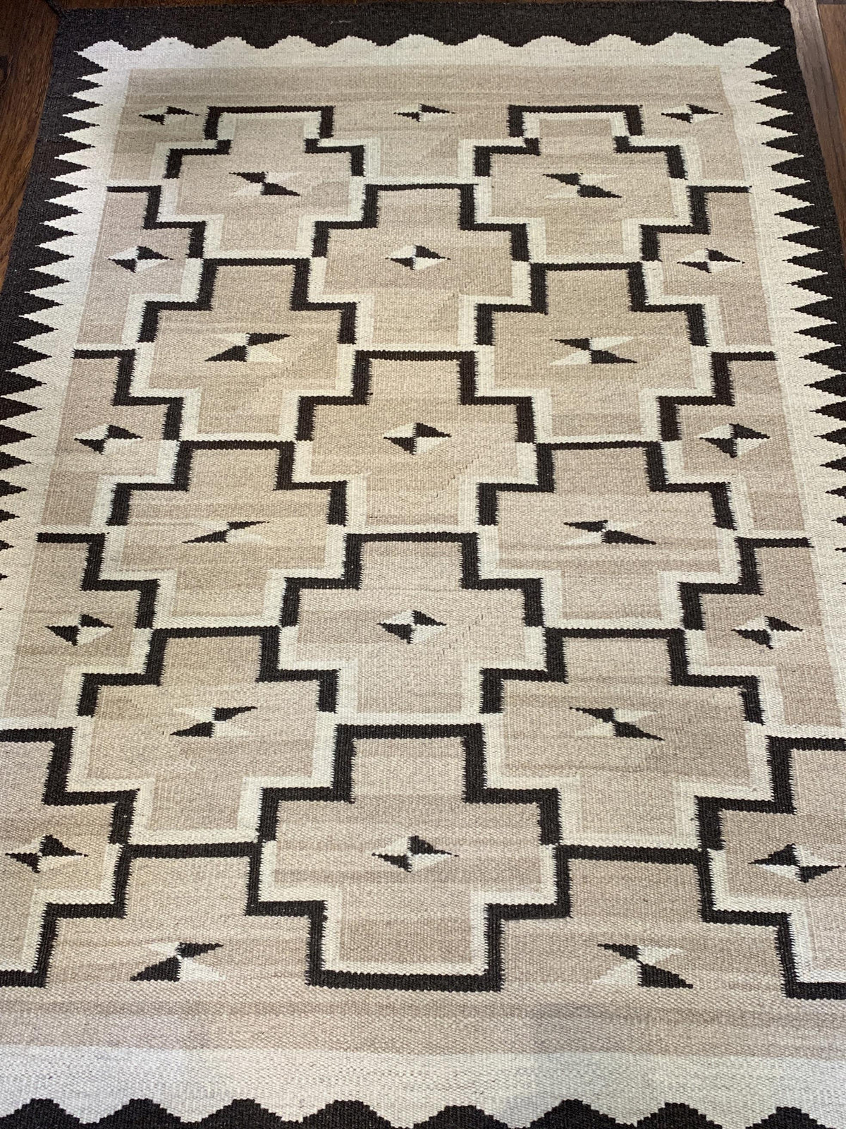 A rectangular woven area rug with a Southwestern geometric pattern in beige, cream, and black, shown flat on a dark wood floor.Atlantica Organics curates antique and handmade rugs, Persian, Afghan, Tibetan, Pakistani, Turkish Oushak, Kilim, Khotan, tribal and contemporary textiles for luxury interiors, homes and real estate. Our collections connect timeless craftsmanship with interior design, furniture, fine art, architecture and modern living. Explore curated rugs, collectible textiles, natural materials,