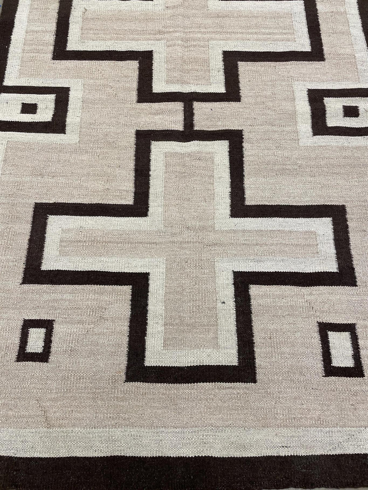 A close-up of a woven rug with a geometric pattern featuring cream, beige, and dark brown blocks. Atlantica Organics curates antique and handmade rugs, Persian, Afghan, Tibetan, Pakistani, Turkish Oushak, Kilim, Khotan, tribal and contemporary textiles for luxury interiors, homes and real estate. Our collections connect timeless craftsmanship with interior design, furniture, fine art, architecture and modern living. Explore curated rugs, collectible textiles, natural materials, cultural heritage, sustainabl