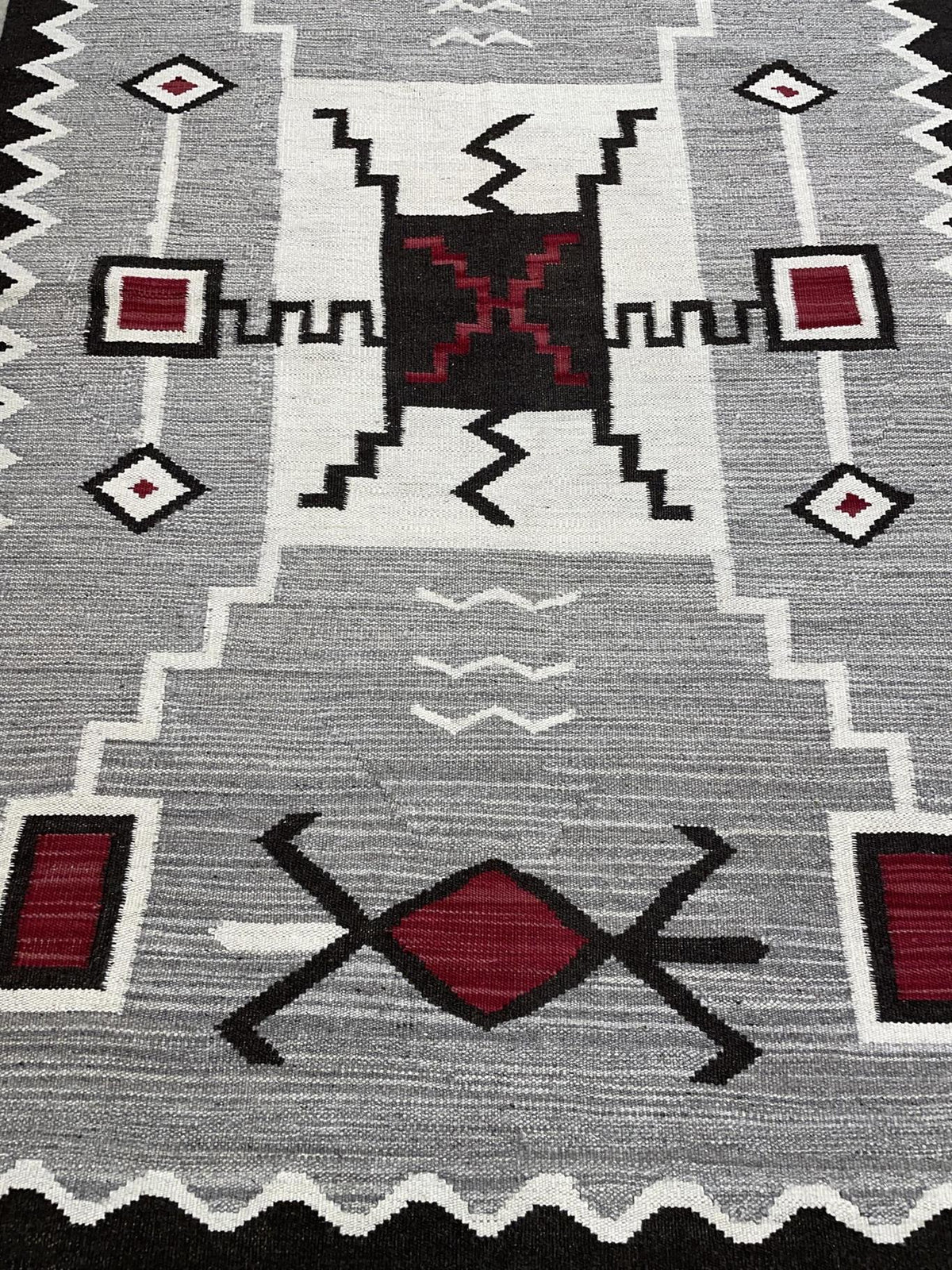A close-up of a woven rug with a Southwestern geometric pattern in gray, white, black, and red. Atlantica Organics curates antique and handmade rugs, Persian, Afghan, Tibetan, Pakistani, Turkish Oushak, Kilim, Khotan, tribal and contemporary textiles for luxury interiors, homes and real estate. Our collections connect timeless craftsmanship with interior design, furniture, fine art, architecture and modern living. Explore curated rugs, collectible textiles, natural materials, cultural heritage, sustainable