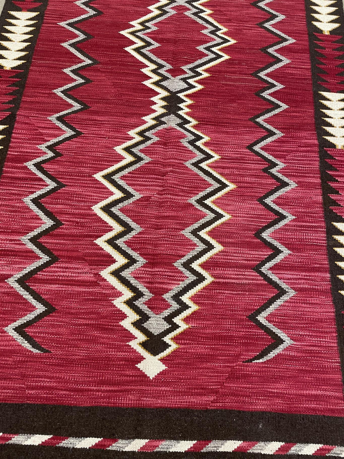 A close-up of a woven rug with a deep red field, featuring bold geometric zigzag and diamond patterns in black, cream, and gray. Atlantica Organics curates antique and handmade rugs, Persian, Afghan, Tibetan, Pakistani, Turkish Oushak, Kilim, Khotan, tribal and contemporary textiles for luxury interiors, homes and real estate. Our collections connect timeless craftsmanship with interior design, furniture, fine art, architecture and modern living. Explore curated rugs, collectible textiles, natural materials