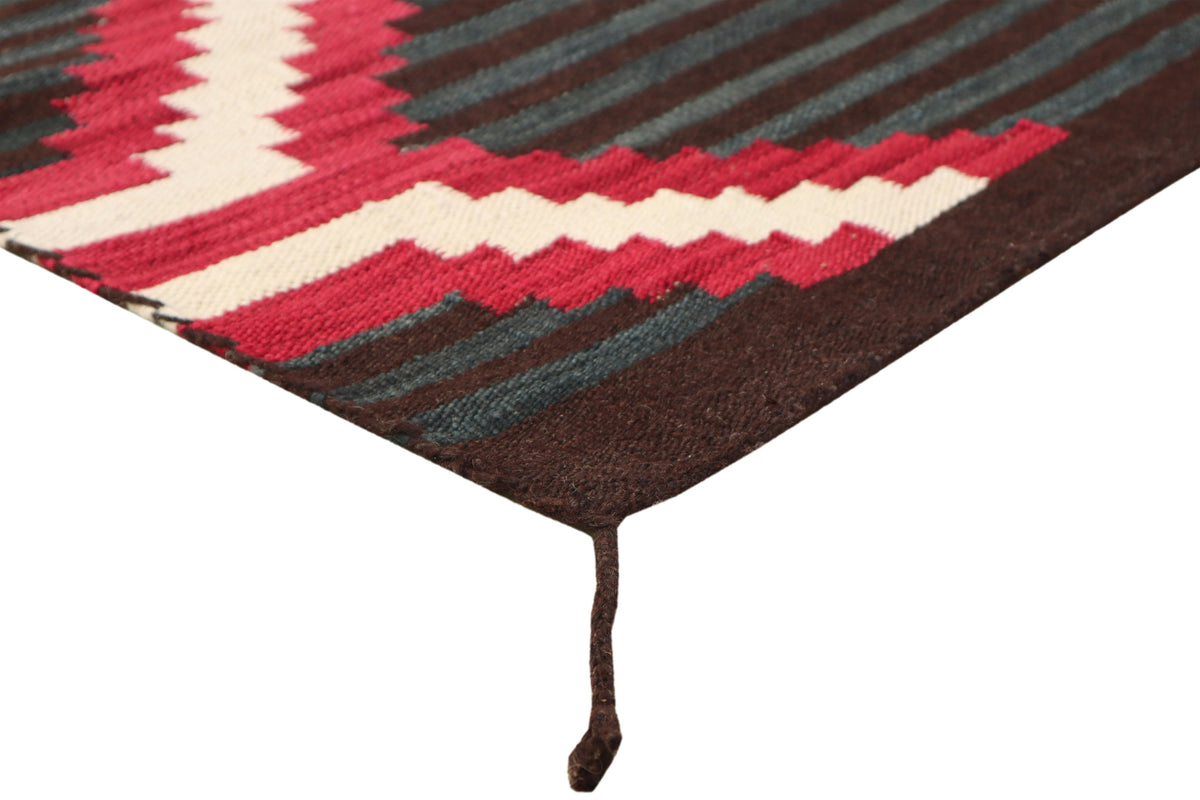 A close-up of a woven rug corner showing a geometric pattern with dark brown, red, cream, and dark teal stripes, finished with a dark brown hanging loop.Atlantica Organics curates antique and handmade rugs, Persian, Afghan, Tibetan, Pakistani, Turkish Oushak, Kilim, Khotan, tribal and contemporary textiles for luxury interiors, homes and real estate. Our collections connect timeless craftsmanship with interior design, furniture, fine art, architecture and modern living. Explore curated rugs, collectible tex