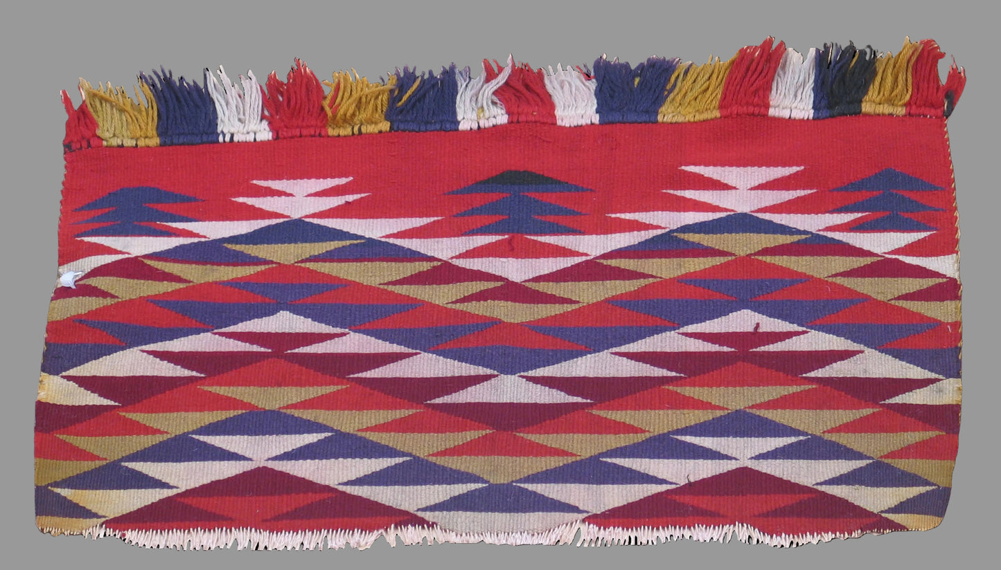 A square woven textile with fringe on the top and bottom edges, featuring a geometric pattern of red, blue, beige, and white triangles and diamonds, shown against a solid black background.
