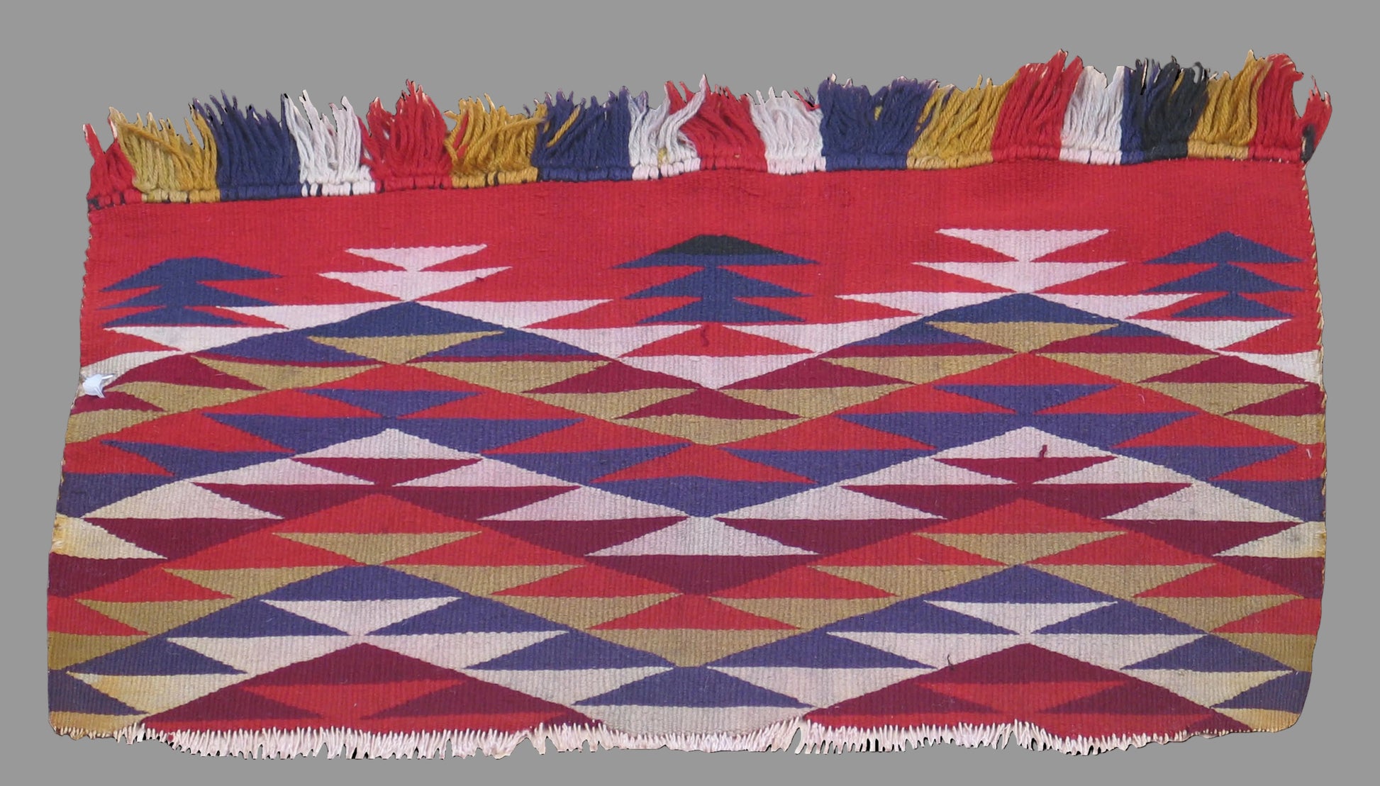 A square woven textile with fringe on the top and bottom edges, featuring a geometric pattern of red, blue, beige, and white triangles and diamonds, shown against a solid black background.