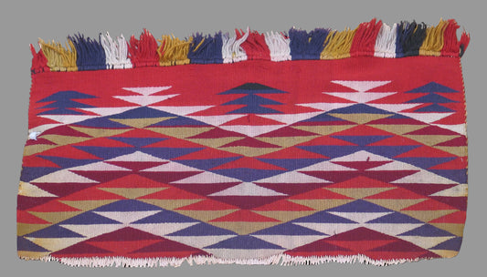A square woven textile with fringe on the top and bottom edges, featuring a geometric pattern of red, blue, beige, and white triangles and diamonds, shown against a solid black background.