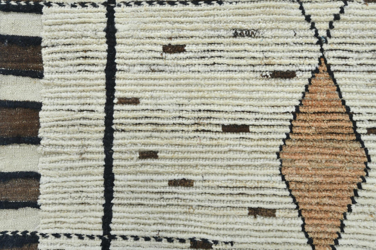 Close-up of a woven rug showing a cream base with black geometric lines, small brown rectangular accents, and a peach diamond motif. 