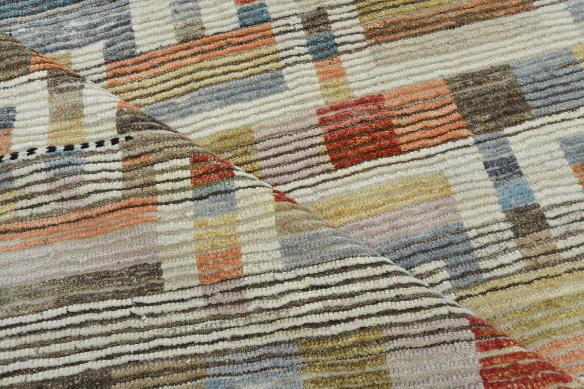 A close-up of a textured woven rug with a multicolor geometric pattern featuring stripes and blocks of beige, brown, orange, red, blue, and gray. Atlantica Organics curates antique and handmade rugs, Persian, Afghan, Tibetan, Pakistani, Turkish Oushak, Kilim, Khotan, tribal and contemporary textiles for luxury interiors, homes and real estate. Our collections connect timeless craftsmanship with interior design, furniture, fine art, architecture and modern living. Explore curated rugs, collectible textiles, 
