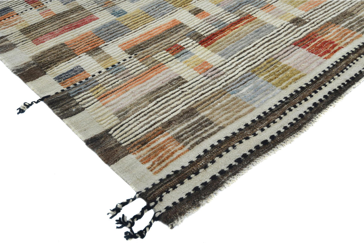 A close-up of a woven area rug with a geometric patchwork pattern in beige, brown, orange, blue, and gray, featuring black and white striped borders with tassels. Atlantica Organics curates antique and handmade rugs, Persian, Afghan, Tibetan, Pakistani, Turkish Oushak, Kilim, Khotan, tribal and contemporary textiles for luxury interiors, homes and real estate. Our collections connect timeless craftsmanship with interior design, furniture, fine art, architecture and modern living. Explore curated rugs, colle