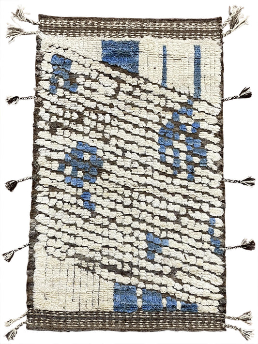 A rectangular handwoven rug with a textured abstract pattern in cream, blue, and dark brown, finished with tassel fringe on all sides.Atlantica Organics curates antique and handmade rugs, Persian, Afghan, Tibetan, Pakistani, Turkish Oushak, Kilim, Khotan, tribal and contemporary textiles for luxury interiors, homes and real estate. Our collections connect timeless craftsmanship with interior design, furniture, fine art, architecture and modern living. Explore curated rugs, collectible textiles, natural mate