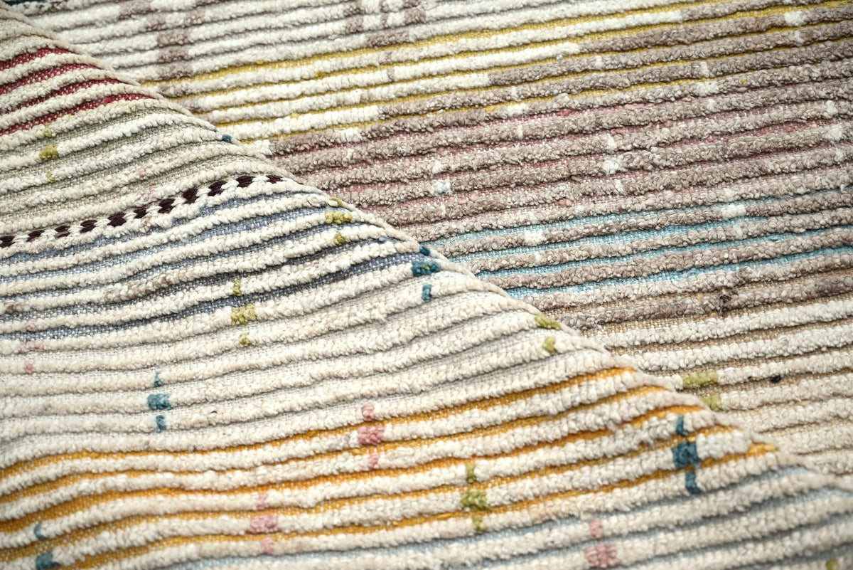 A close-up of a textured woven rug with horizontal stripes in cream, beige, brown, muted blue, and small multicolor accents. Atlantica Organics curates antique and handmade rugs, Persian, Afghan, Tibetan, Pakistani, Turkish Oushak, Kilim, Khotan, tribal and contemporary textiles for luxury interiors, homes and real estate. Our collections connect timeless craftsmanship with interior design, furniture, fine art, architecture and modern living. Explore curated rugs, collectible textiles, natural materials, cu