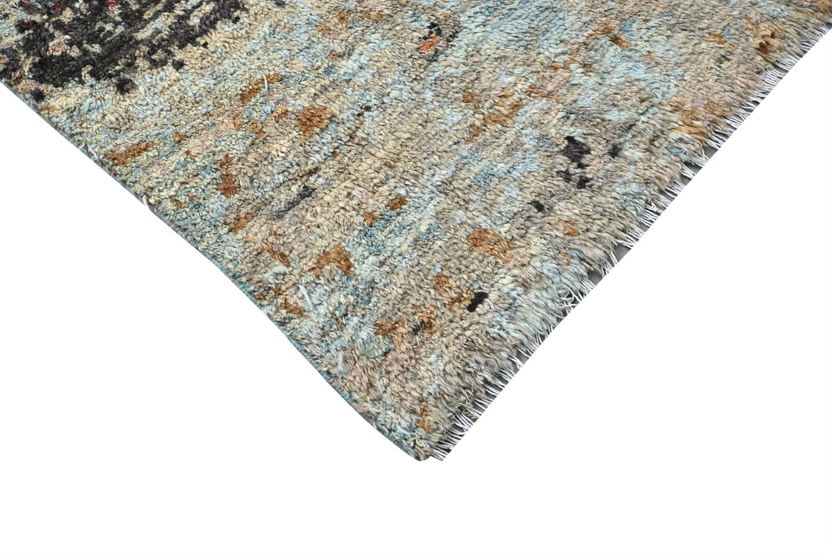 A close-up corner view of a textured area rug with a distressed, abstract pattern in light blue, beige, brown, and black, featuring a fringed edge on a white background. Atlantica Organics curates antique and handmade rugs, Persian, Afghan, Tibetan, Pakistani, Turkish Oushak, Kilim, Khotan, tribal and contemporary textiles for luxury interiors, homes and real estate. Our collections connect timeless craftsmanship with interior design, furniture, fine art, architecture and modern living. Explore curated rugs