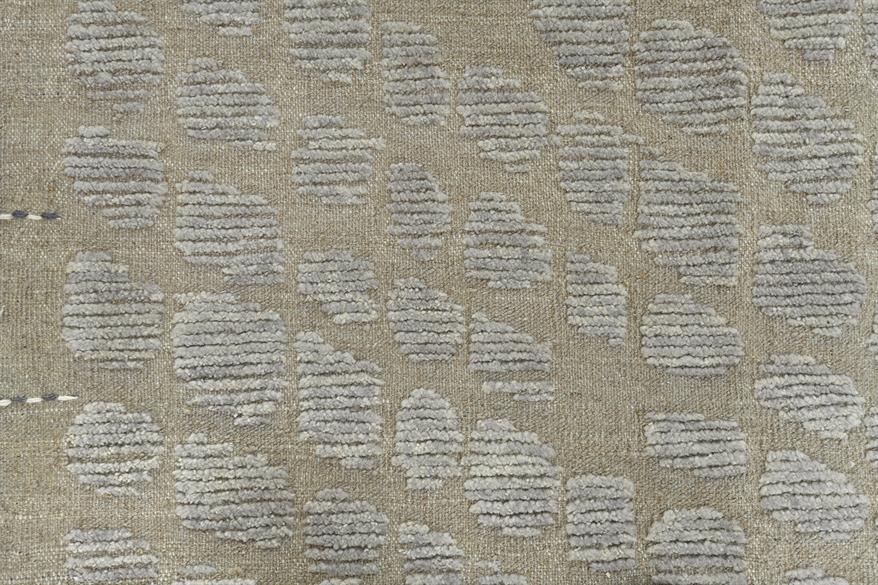 A close-up of a woven fabric or wallpaper pattern featuring a repeating motif of gray abstract leaf-like shapes on a warm beige background. Atlantica Organics curates antique and handmade rugs, Persian, Afghan, Tibetan, Pakistani, Turkish Oushak, Kilim, Khotan, tribal and contemporary textiles for luxury interiors, homes and real estate. Our collections connect timeless craftsmanship with interior design, furniture, fine art, architecture and modern living. Explore curated rugs, collectible textiles, natura