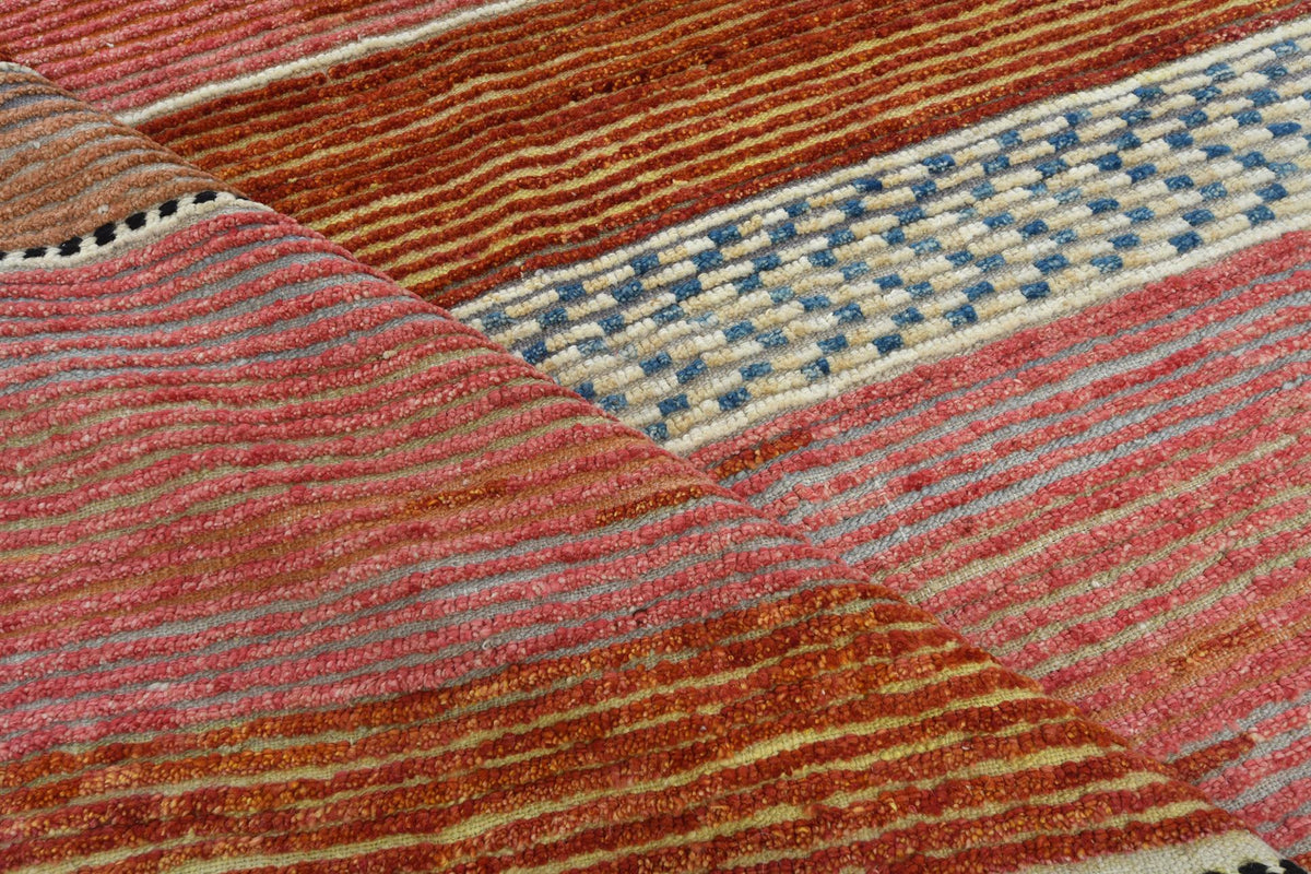 A close-up of a textured woven rug with horizontal stripes in rust orange, pink, beige, and a blue geometric patterned band. Atlantica Organics curates antique and handmade rugs, Persian, Afghan, Tibetan, Pakistani, Turkish Oushak, Kilim, Khotan, tribal and contemporary textiles for luxury interiors, homes and real estate. Our collections connect timeless craftsmanship with interior design, furniture, fine art, architecture and modern living. Explore curated rugs, collectible textiles, natural materials, cu
