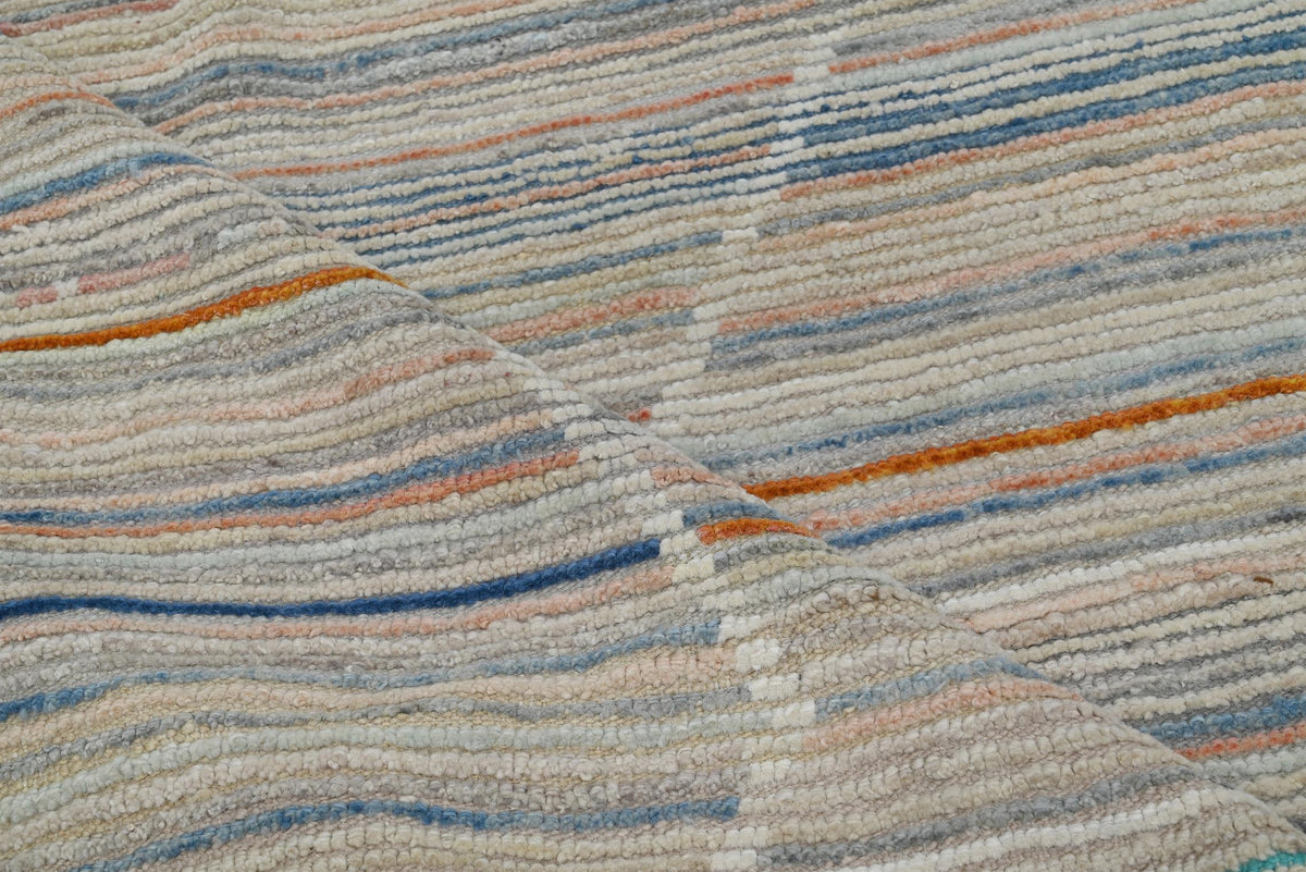 A close-up of a textured woven rug with horizontal stripes in beige, cream, light blue, orange, and gray. Atlantica Organics curates antique and handmade rugs, Persian, Afghan, Tibetan, Pakistani, Turkish Oushak, Kilim, Khotan, tribal and contemporary textiles for luxury interiors, homes and real estate. Our collections connect timeless craftsmanship with interior design, furniture, fine art, architecture and modern living. Explore curated rugs, collectible textiles, natural materials, cultural heritage, su