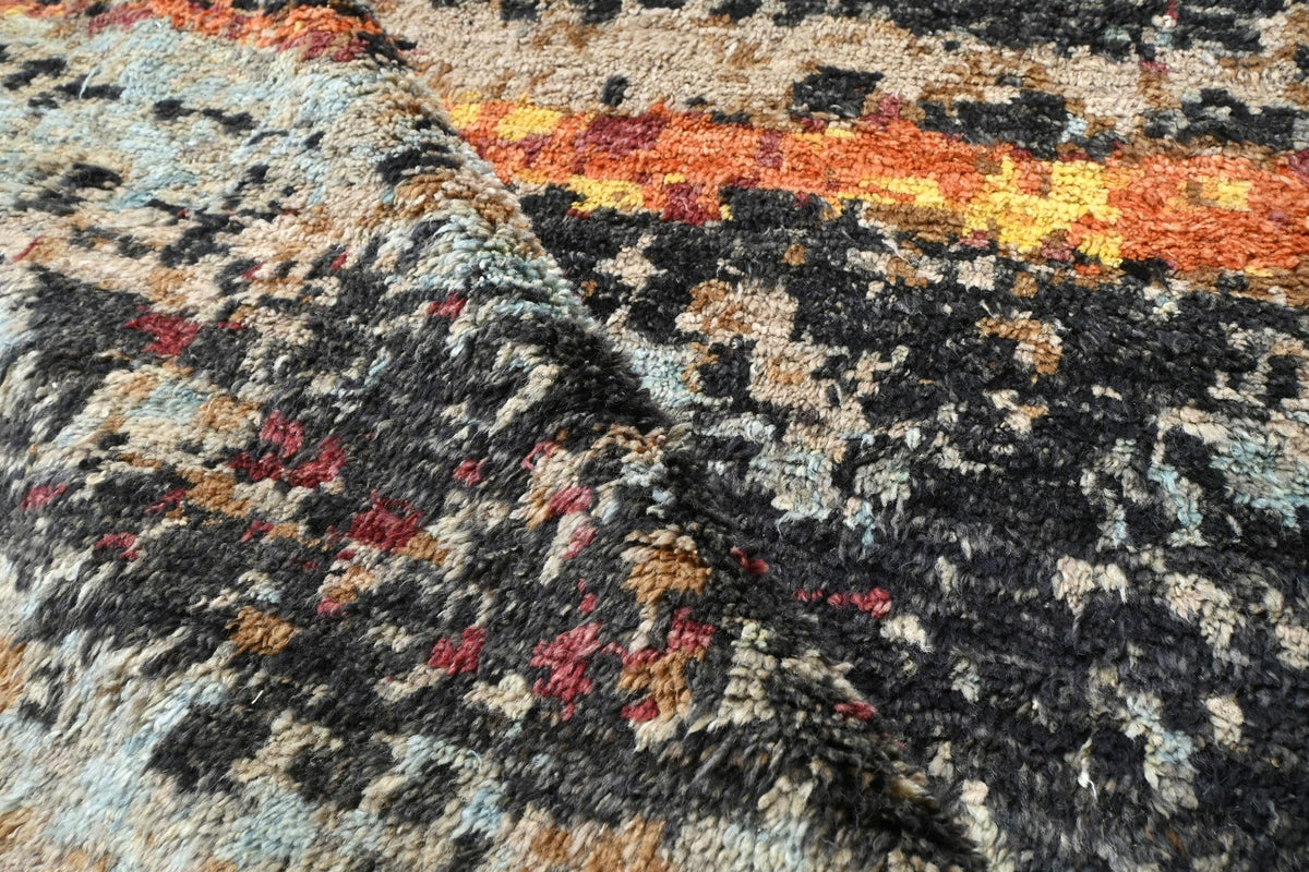 Close-up of a textured, shaggy area rug with a mottled abstract pattern in black, gray, beige, orange, yellow, and red. Atlantica Organics curates antique and handmade rugs, Persian, Afghan, Tibetan, Pakistani, Turkish Oushak, Kilim, Khotan, tribal and contemporary textiles for luxury interiors, homes and real estate. Our collections connect timeless craftsmanship with interior design, furniture, fine art, architecture and modern living. Explore curated rugs, collectible textiles, natural materials, cultura