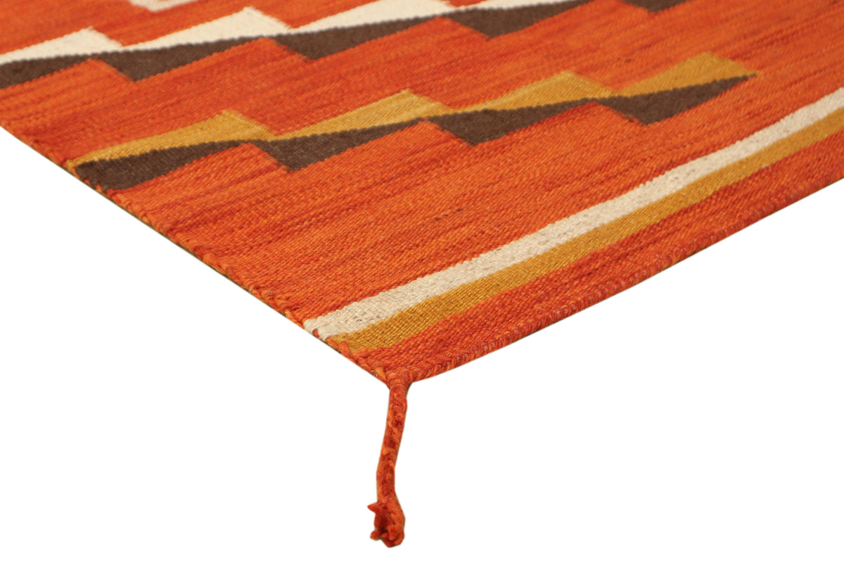 A close-up of a woven textile corner showing a geometric pattern in orange, brown, cream, and mustard yellow with a matching orange tassel fringe on a white background.Atlantica Organics curates antique and handmade rugs, Persian, Afghan, Tibetan, Pakistani, Turkish Oushak, Kilim, Khotan, tribal and contemporary textiles for luxury interiors, homes and real estate. Our collections connect timeless craftsmanship with interior design, furniture, fine art, architecture and modern living. Explore curated rugs, 