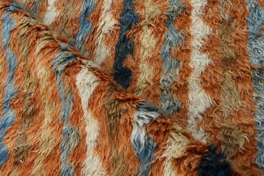 A close-up of a shaggy, textured faux fur or wool fabric with vertical stripes in rust orange, cream, slate blue, and charcoal gray. Atlantica Organics curates antique and handmade rugs, Persian, Afghan, Tibetan, Pakistani, Turkish Oushak, Kilim, Khotan, tribal and contemporary textiles for luxury interiors, homes and real estate. Our collections connect timeless craftsmanship with interior design, furniture, fine art, architecture and modern living. Explore curated rugs, collectible textiles, natural mater