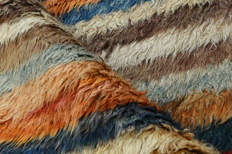 A close-up of a shaggy, textured faux fur or sheepskin material with horizontal stripes in cream, brown, blue, orange, and gray. Atlantica Organics curates antique and handmade rugs, Persian, Afghan, Tibetan, Pakistani, Turkish Oushak, Kilim, Khotan, tribal and contemporary textiles for luxury interiors, homes and real estate. Our collections connect timeless craftsmanship with interior design, furniture, fine art, architecture and modern living. Explore curated rugs, collectible textiles, natural materials