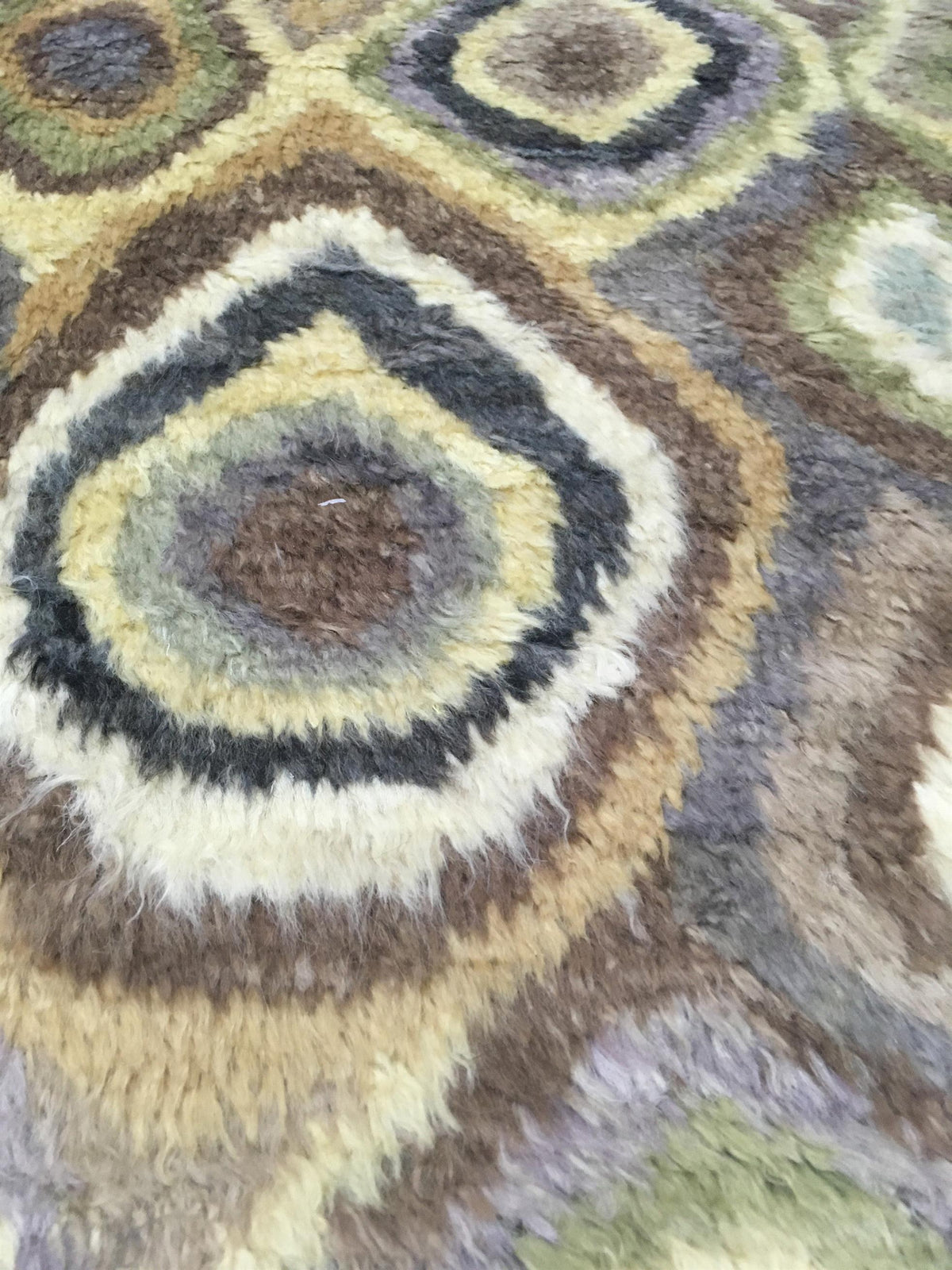 Close-up of a shaggy area rug with a geometric medallion pattern in brown, beige, gray, black, and muted green. Atlantica Organics curates antique and handmade rugs, Persian, Afghan, Tibetan, Pakistani, Turkish Oushak, Kilim, Khotan, tribal and contemporary textiles for luxury interiors, homes and real estate. Our collections connect timeless craftsmanship with interior design, furniture, fine art, architecture and modern living. Explore curated rugs, collectible textiles, natural materials, cultural herita