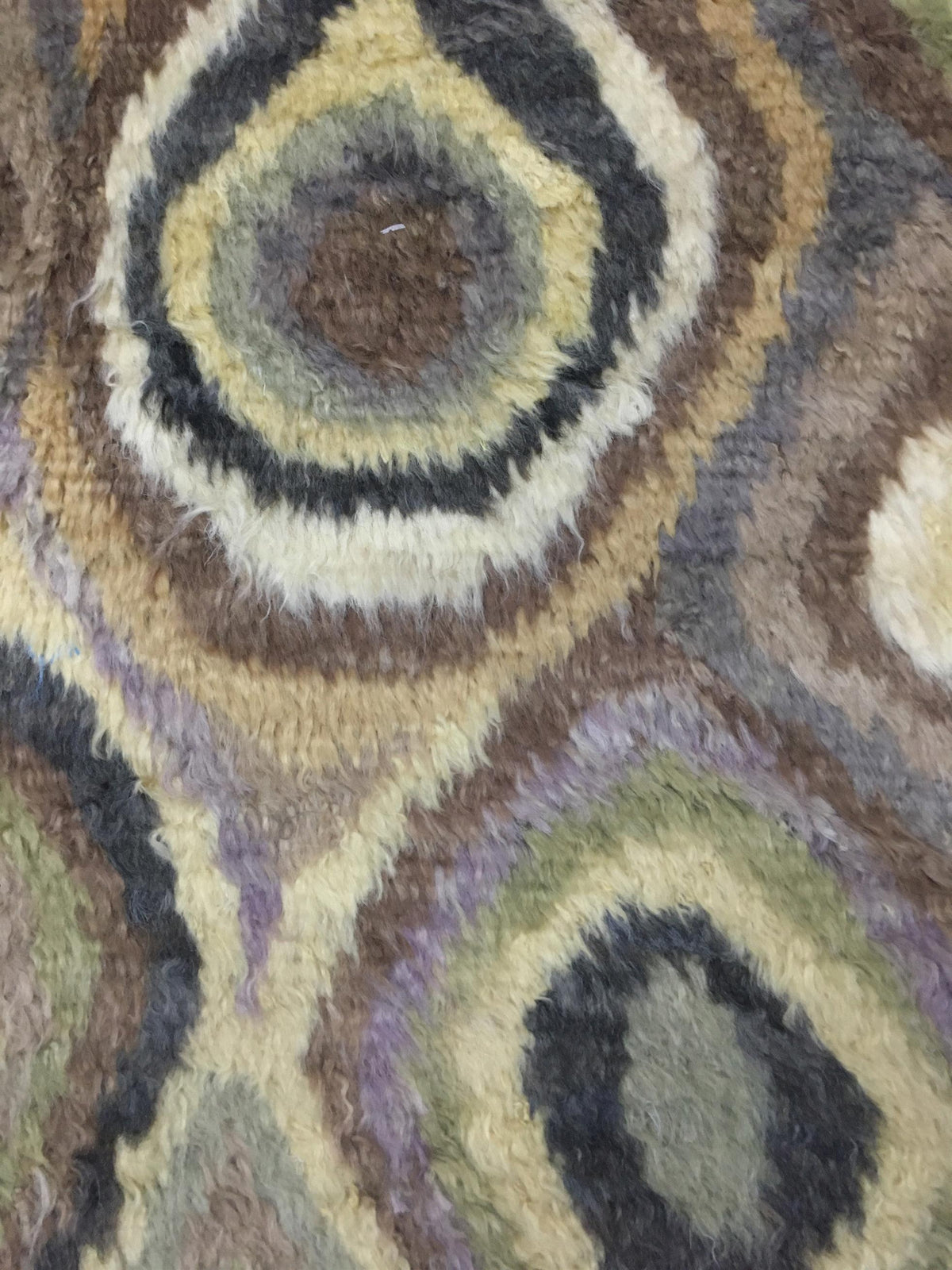 A close-up of a shaggy area rug featuring a retro concentric oval pattern in muted tones of brown, beige, gray, olive green, and lavender. Atlantica Organics curates antique and handmade rugs, Persian, Afghan, Tibetan, Pakistani, Turkish Oushak, Kilim, Khotan, tribal and contemporary textiles for luxury interiors, homes and real estate. Our collections connect timeless craftsmanship with interior design, furniture, fine art, architecture and modern living. Explore curated rugs, collectible textiles, natural