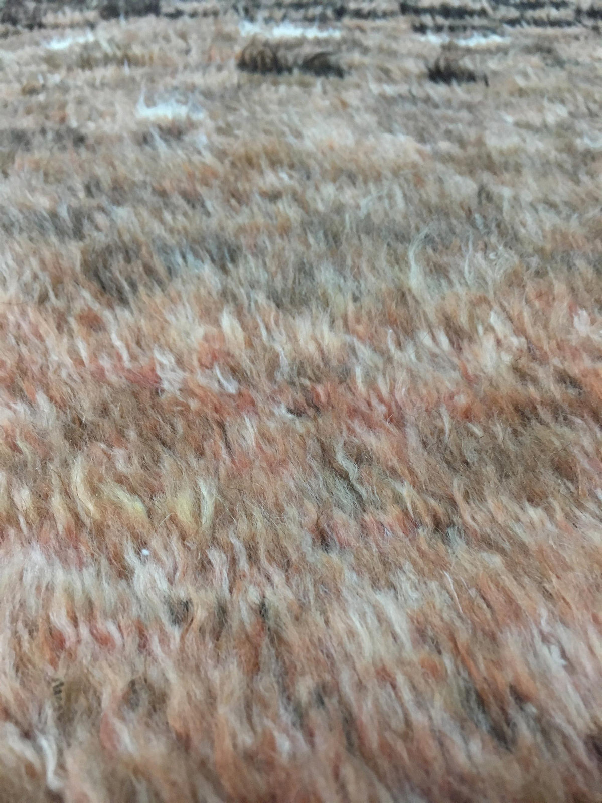 A close-up texture shot of a shaggy, curly wool or fur-like material in a mottled blend of beige, brown, peach, and cream tones. Atlantica Organics curates antique and handmade rugs, Persian, Afghan, Tibetan, Pakistani, Turkish Oushak, Kilim, Khotan, tribal and contemporary textiles for luxury interiors, homes and real estate. Our collections connect timeless craftsmanship with interior design, furniture, fine art, architecture and modern living. Explore curated rugs, collectible textiles, natural materials