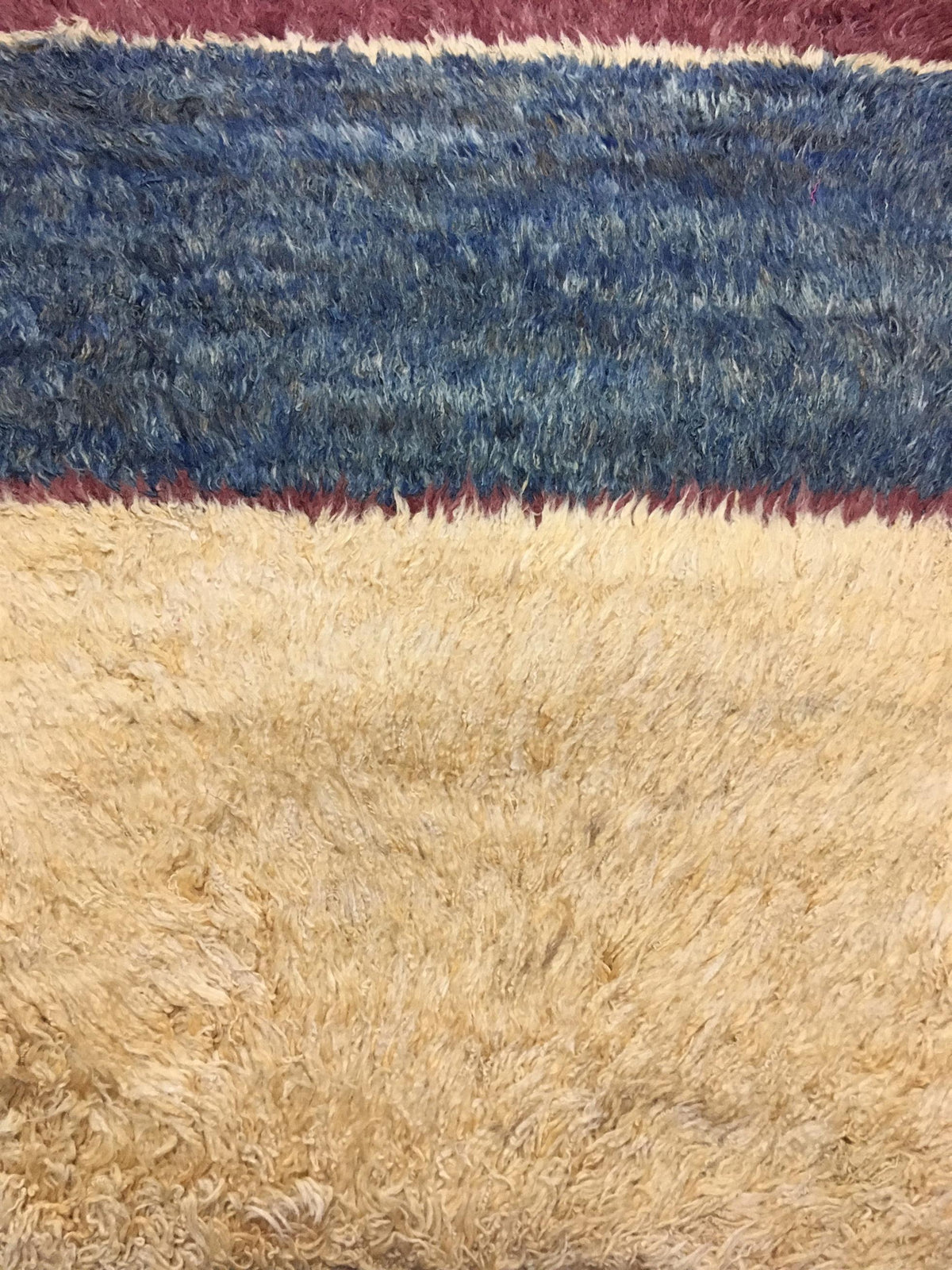 A close-up of a shaggy rug with horizontal stripes, featuring a large beige section at the bottom, a medium blue section in the middle, and a burgundy stripe near the top. Atlantica Organics curates antique and handmade rugs, Persian, Afghan, Tibetan, Pakistani, Turkish Oushak, Kilim, Khotan, tribal and contemporary textiles for luxury interiors, homes and real estate. Our collections connect timeless craftsmanship with interior design, furniture, fine art, architecture and modern living. Explore curated ru