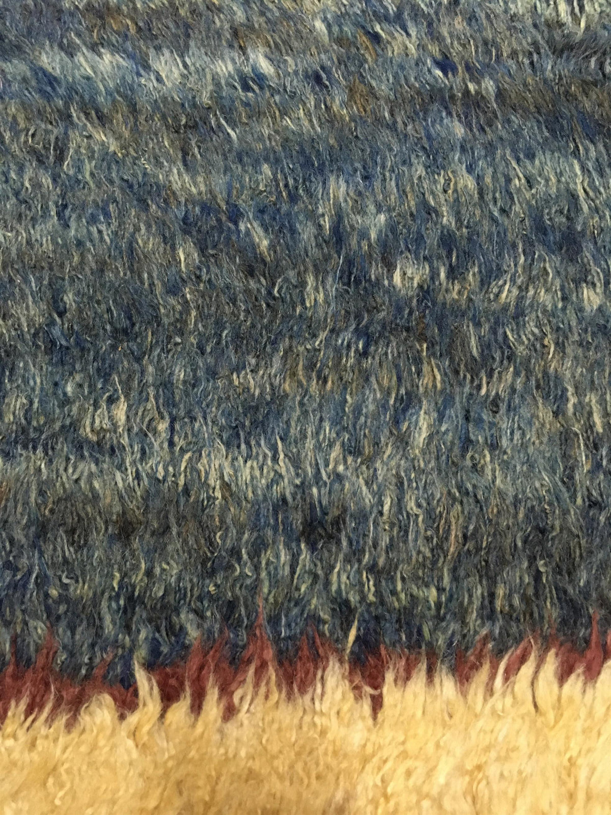 Close-up of a shaggy faux fur or textured fabric surface, showing a mottled blue-gray upper section and a warm cream lower section with a thin rust-brown fringe. Atlantica Organics curates antique and handmade rugs, Persian, Afghan, Tibetan, Pakistani, Turkish Oushak, Kilim, Khotan, tribal and contemporary textiles for luxury interiors, homes and real estate. Our collections connect timeless craftsmanship with interior design, furniture, fine art, architecture and modern living. Explore curated rugs, collec