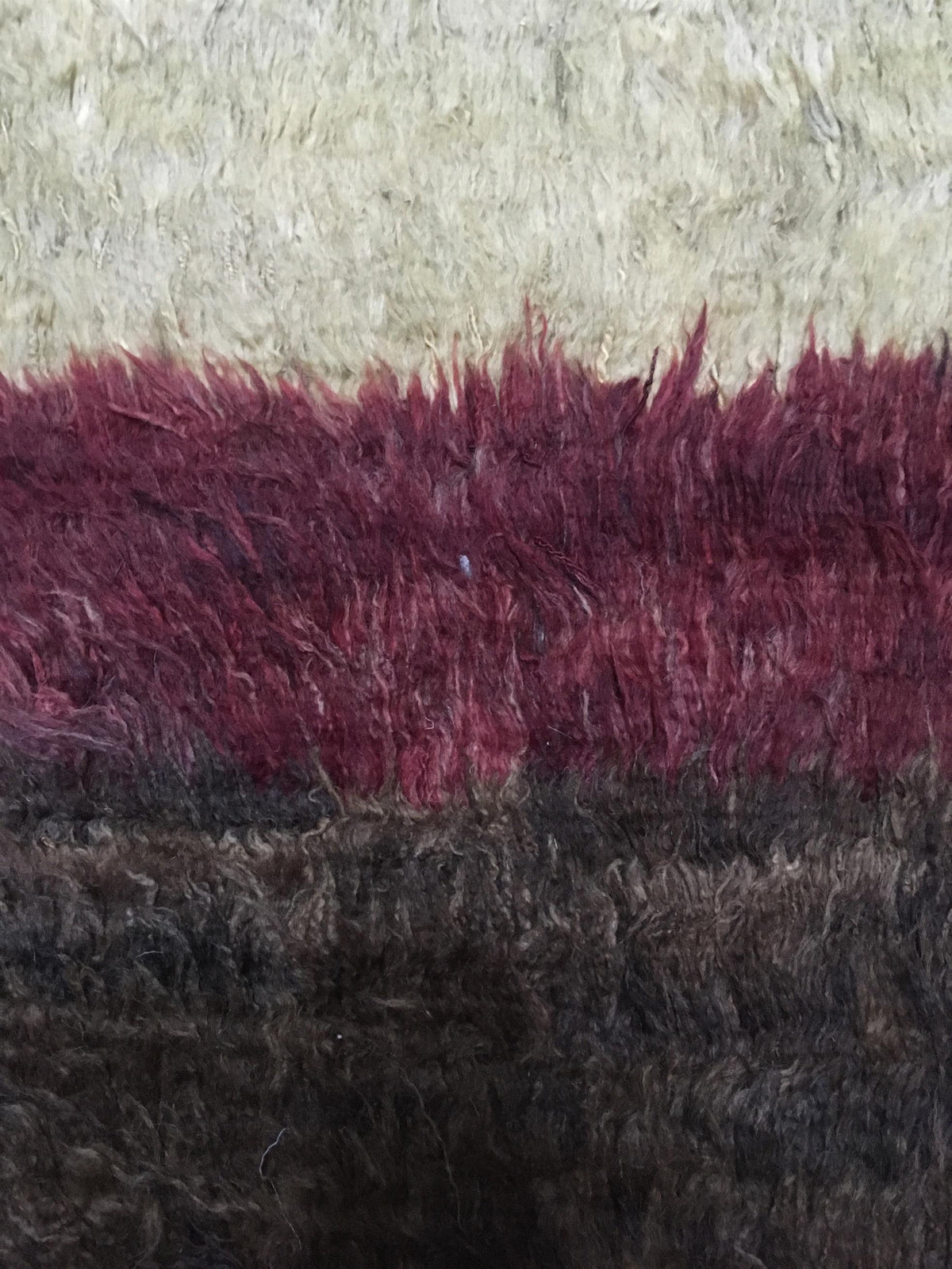 A close-up of a shaggy, textured rug or fabric with horizontal color bands: a cream top section, a wide deep burgundy middle band, and a dark brown bottom section. Atlantica Organics curates antique and handmade rugs, Persian, Afghan, Tibetan, Pakistani, Turkish Oushak, Kilim, Khotan, tribal and contemporary textiles for luxury interiors, homes and real estate. Our collections connect timeless craftsmanship with interior design, furniture, fine art, architecture and modern living. Explore curated rugs, coll