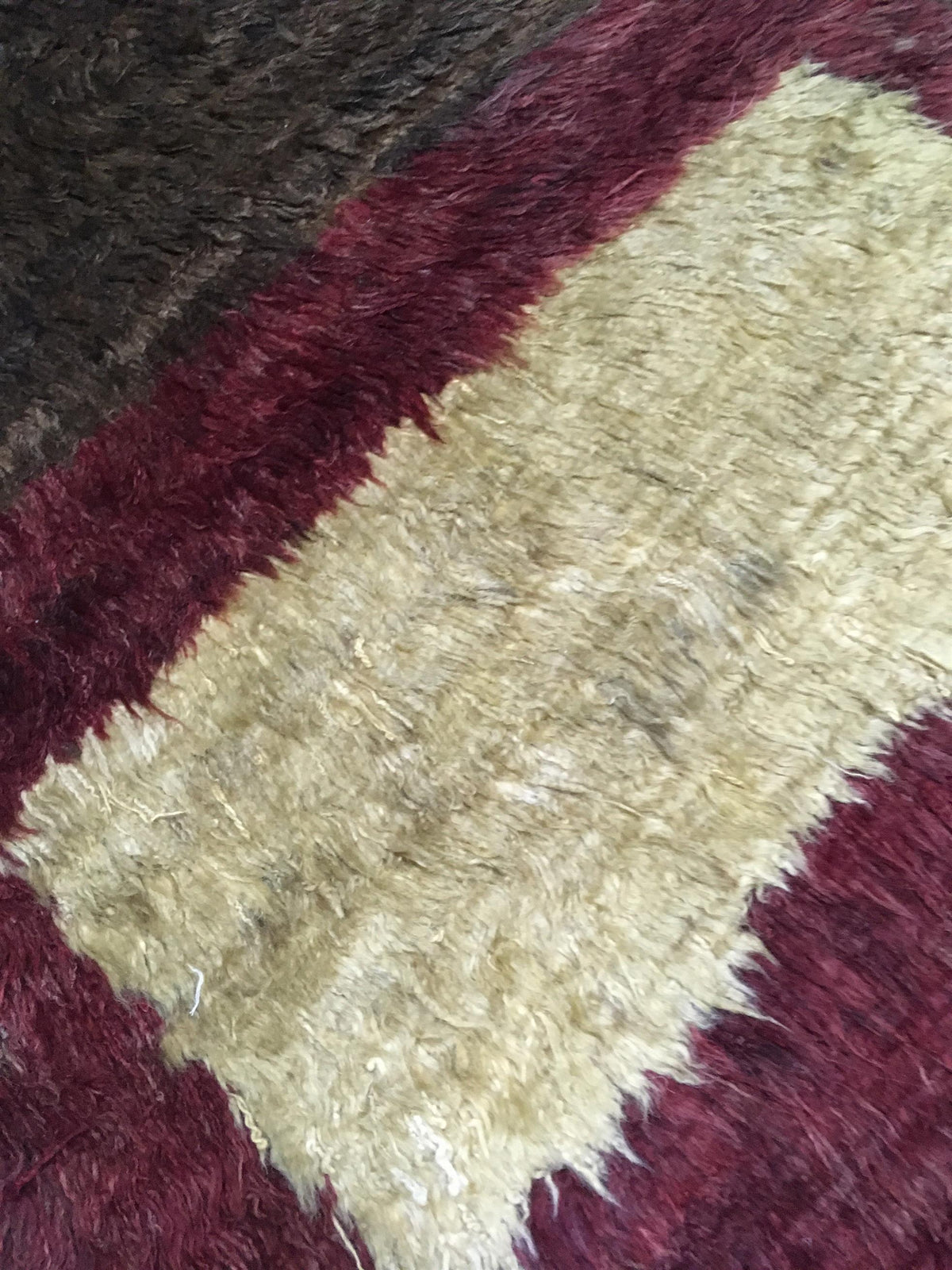 A close-up of a shaggy area rug with a geometric pattern, featuring a central light beige block bordered by deep burgundy and a dark brown section in the upper left. Atlantica Organics curates antique and handmade rugs, Persian, Afghan, Tibetan, Pakistani, Turkish Oushak, Kilim, Khotan, tribal and contemporary textiles for luxury interiors, homes and real estate. Our collections connect timeless craftsmanship with interior design, furniture, fine art, architecture and modern living. Explore curated rugs, co
