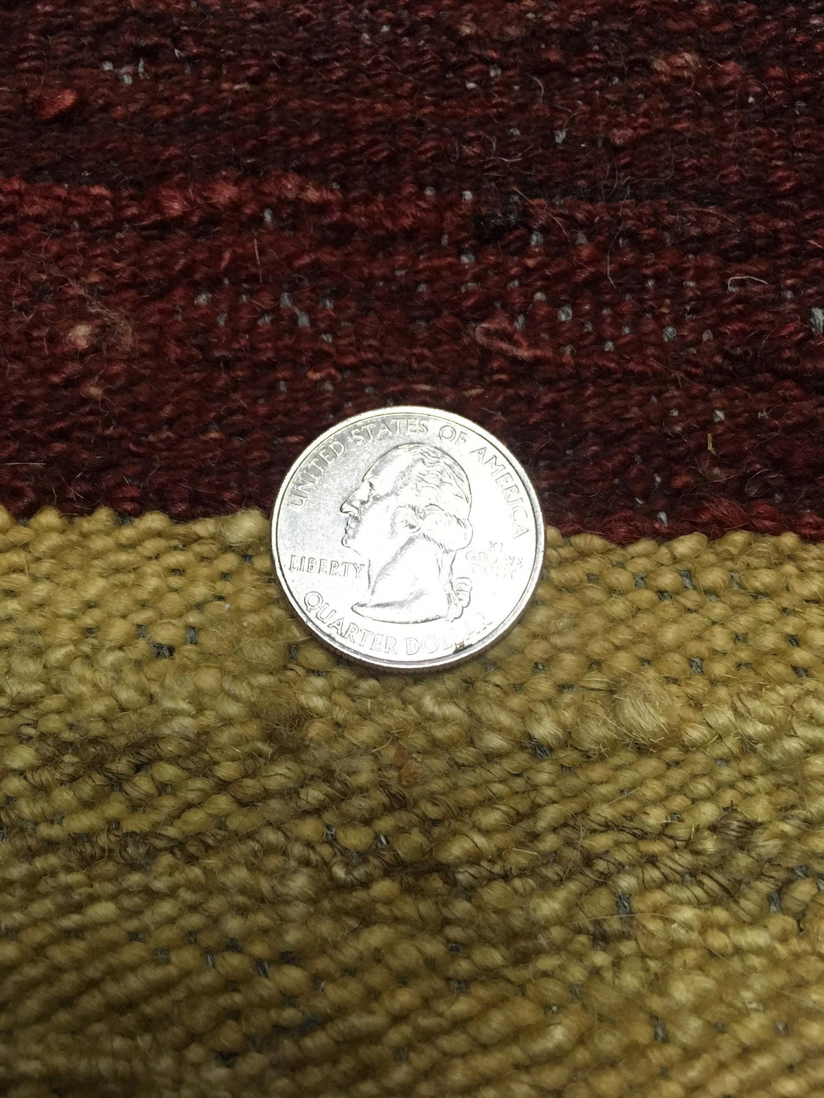 A silver US quarter coin placed on a textured woven rug with horizontal stripes of dark red on top and olive green below. Atlantica Organics curates antique and handmade rugs, Persian, Afghan, Tibetan, Pakistani, Turkish Oushak, Kilim, Khotan, tribal and contemporary textiles for luxury interiors, homes and real estate. Our collections connect timeless craftsmanship with interior design, furniture, fine art, architecture and modern living. Explore curated rugs, collectible textiles, natural materials, cultu