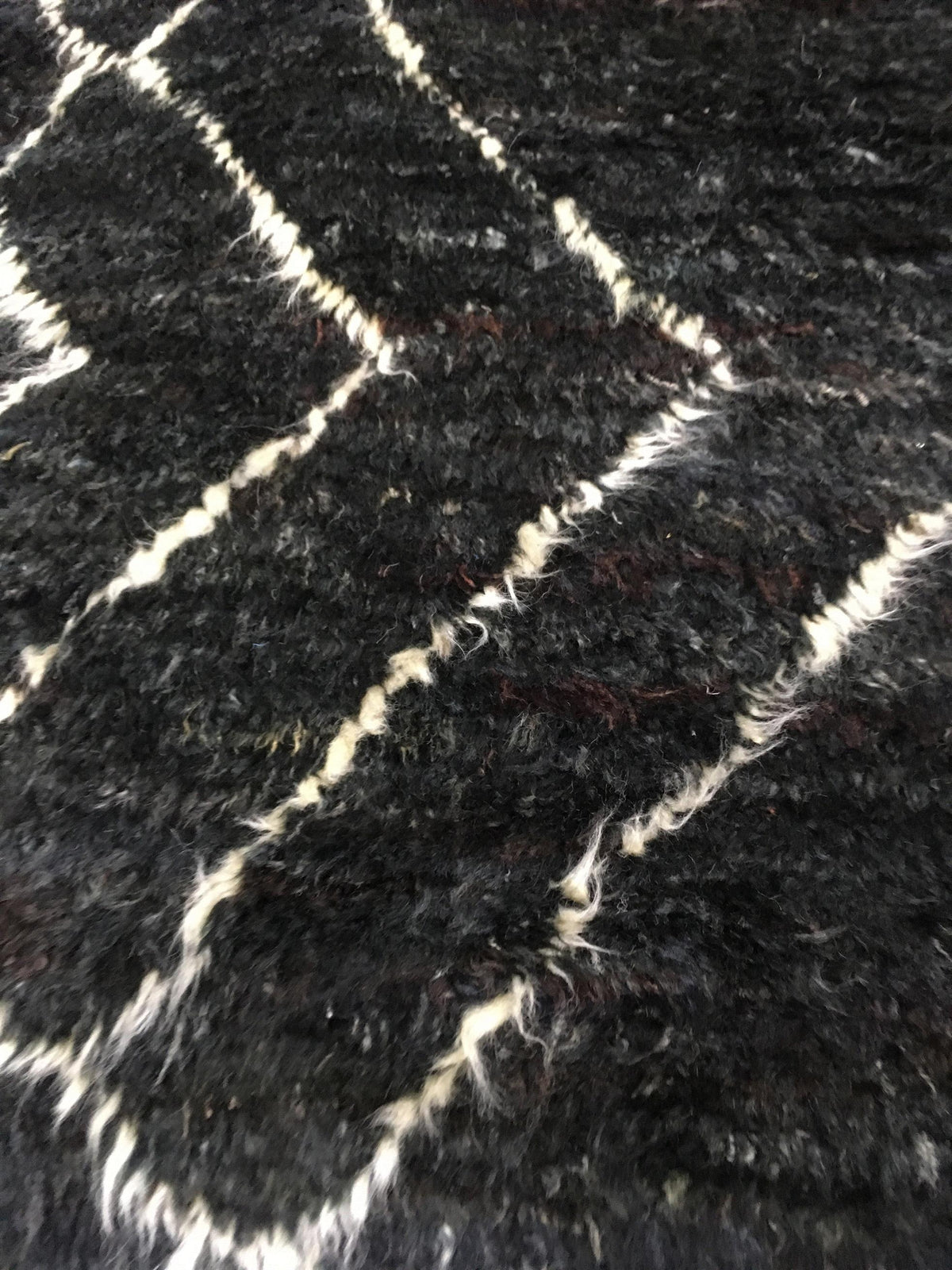 A close-up of a black shaggy or faux-fur textured surface with irregular off-white stitched or woven lines forming a geometric pattern. Atlantica Organics curates antique and handmade rugs, Persian, Afghan, Tibetan, Pakistani, Turkish Oushak, Kilim, Khotan, tribal and contemporary textiles for luxury interiors, homes and real estate. Our collections connect timeless craftsmanship with interior design, furniture, fine art, architecture and modern living. Explore curated rugs, collectible textiles, natural ma