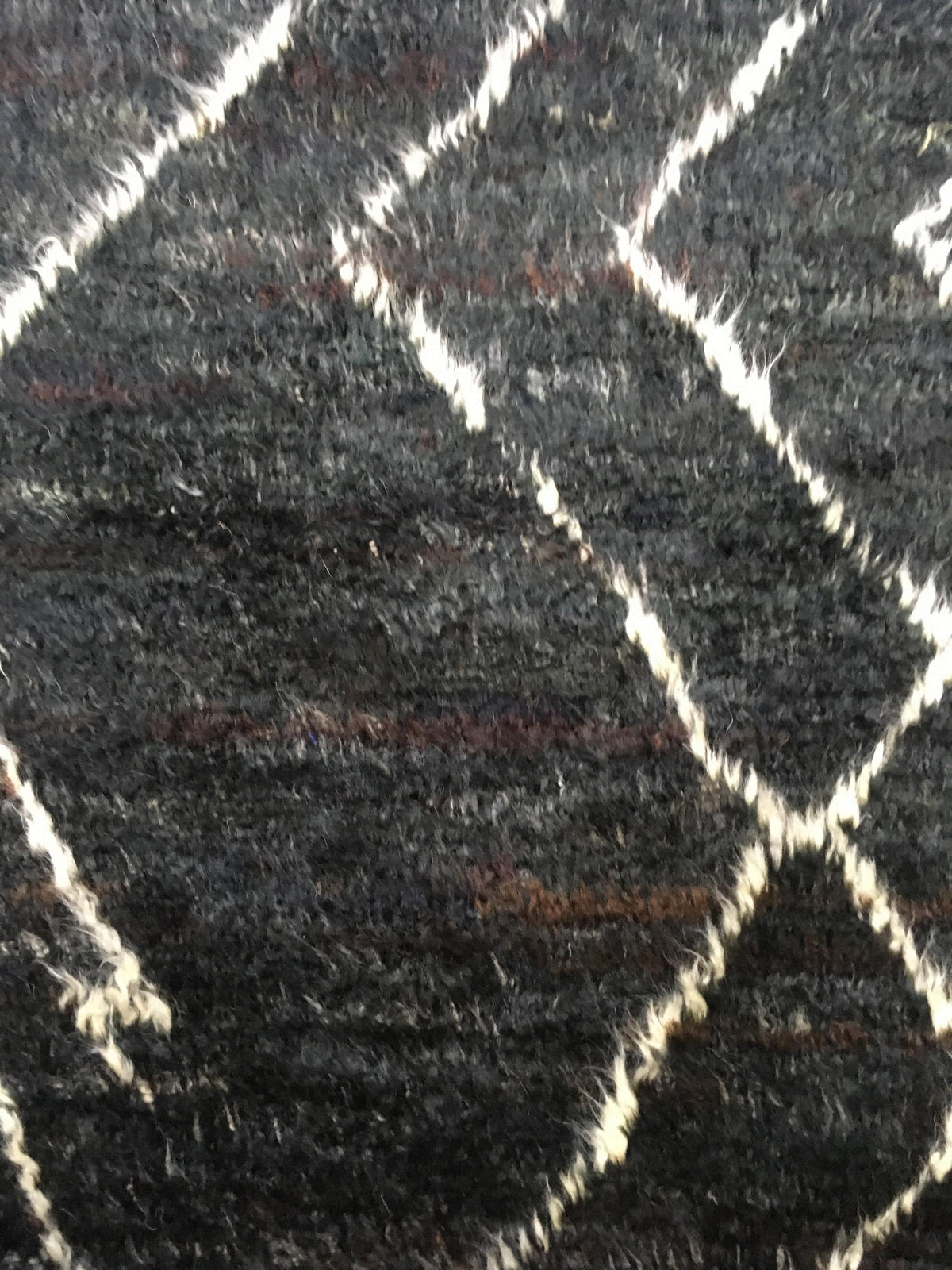 A close-up of a dark charcoal gray textured rug with a cream-colored geometric diamond pattern. Atlantica Organics curates antique and handmade rugs, Persian, Afghan, Tibetan, Pakistani, Turkish Oushak, Kilim, Khotan, tribal and contemporary textiles for luxury interiors, homes and real estate. Our collections connect timeless craftsmanship with interior design, furniture, fine art, architecture and modern living. Explore curated rugs, collectible textiles, natural materials, cultural heritage, sustainable 