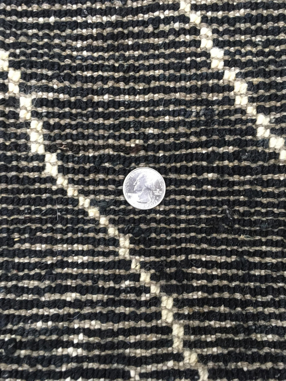 Close-up of a woven textile surface in black and off-white with a small silver coin placed on top for scale. Atlantica Organics curates antique and handmade rugs, Persian, Afghan, Tibetan, Pakistani, Turkish Oushak, Kilim, Khotan, tribal and contemporary textiles for luxury interiors, homes and real estate. Our collections connect timeless craftsmanship with interior design, furniture, fine art, architecture and modern living. Explore curated rugs, collectible textiles, natural materials, cultural heritage,