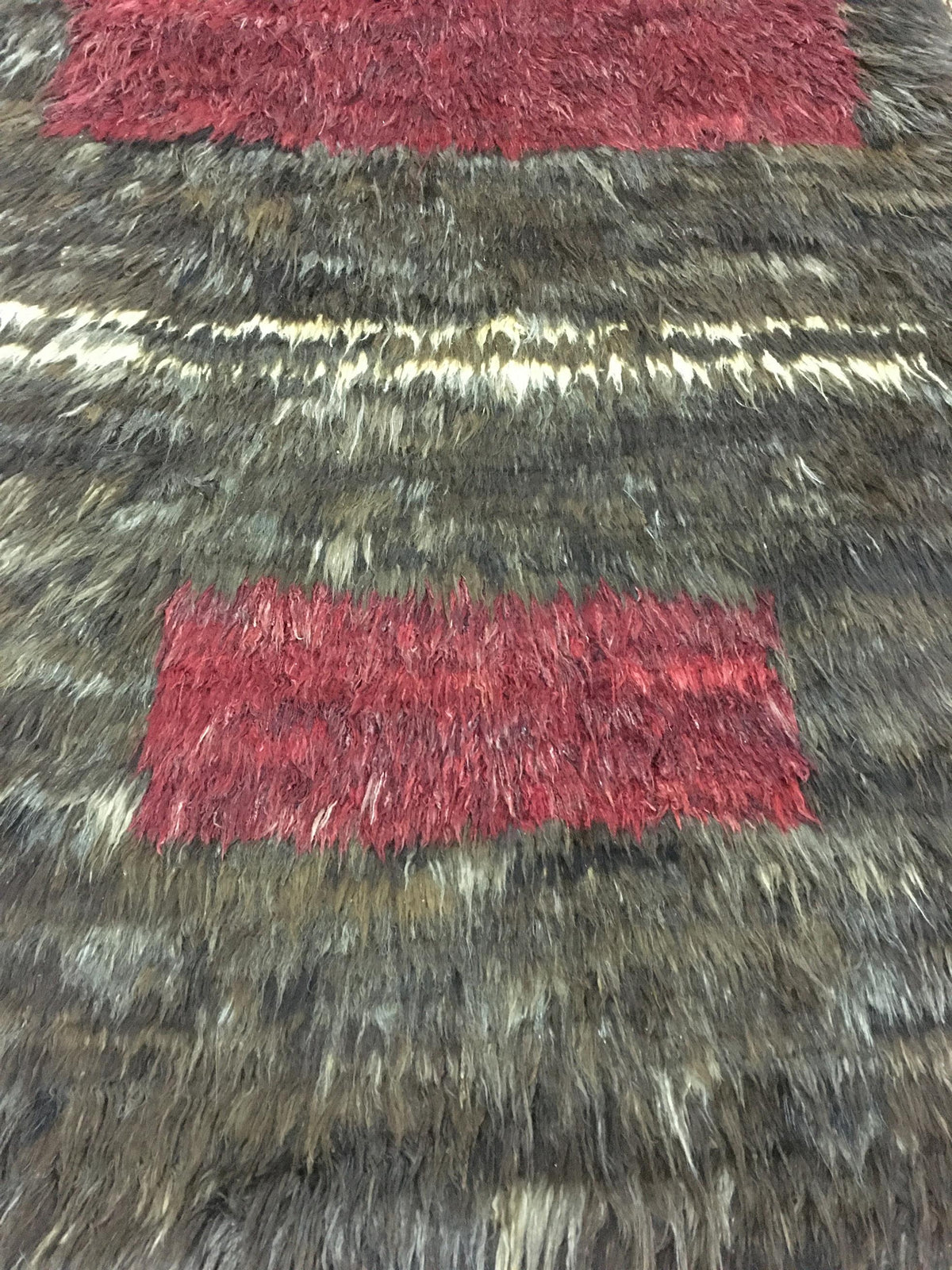A close-up of a shaggy area rug with a textured, mottled pattern in dark brown, gray, and cream, featuring two rectangular blocks of deep red shag on either side of a horizontal band. Atlantica Organics curates antique and handmade rugs, Persian, Afghan, Tibetan, Pakistani, Turkish Oushak, Kilim, Khotan, tribal and contemporary textiles for luxury interiors, homes and real estate. Our collections connect timeless craftsmanship with interior design, furniture, fine art, architecture and modern living. Explor