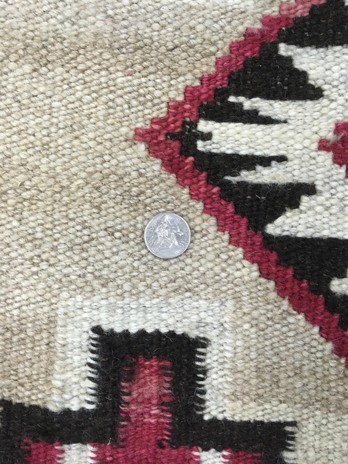 Close-up of a woven rug or textile showing a cream base with black and dark red geometric patterns, with a coin placed in the center for scale. Atlantica Organics curates antique and handmade rugs, Persian, Afghan, Tibetan, Pakistani, Turkish Oushak, Kilim, Khotan, tribal and contemporary textiles for luxury interiors, homes and real estate. Our collections connect timeless craftsmanship with interior design, furniture, fine art, architecture and modern living. Explore curated rugs, collectible textiles, na