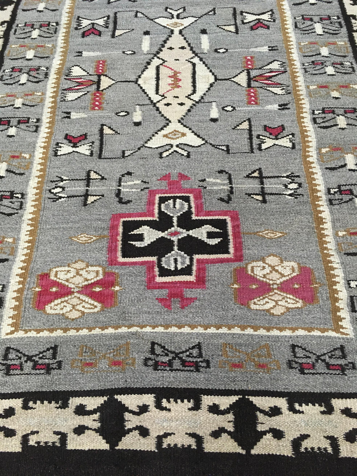 A close-up of a woven geometric rug with a gray field, featuring a central black and pink cross motif, cream and black tribal symbols, and a black border at the bottom. Atlantica Organics curates antique and handmade rugs, Persian, Afghan, Tibetan, Pakistani, Turkish Oushak, Kilim, Khotan, tribal and contemporary textiles for luxury interiors, homes and real estate. Our collections connect timeless craftsmanship with interior design, furniture, fine art, architecture and modern living. Explore curated rugs,