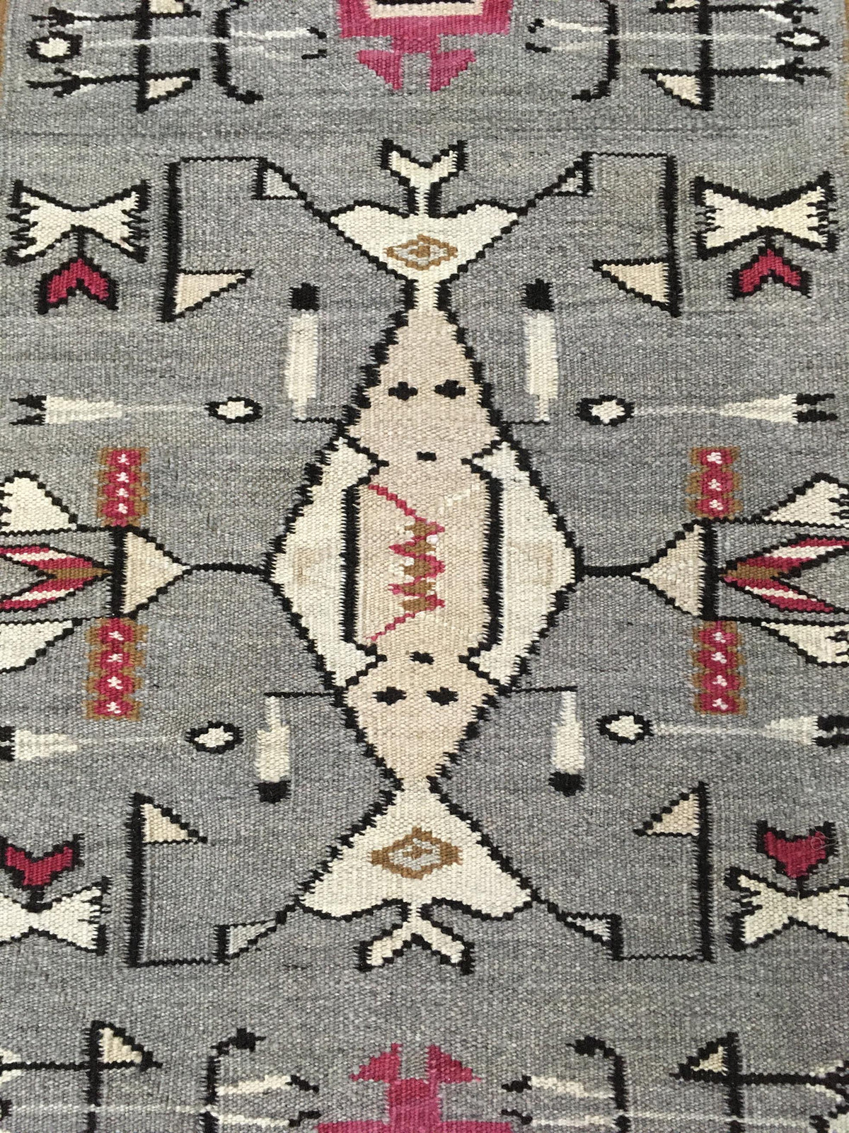 A close-up of a woven rug with a gray background and a central diamond motif in cream, black, red, and pink, surrounded by geometric tribal patterns. Atlantica Organics curates antique and handmade rugs, Persian, Afghan, Tibetan, Pakistani, Turkish Oushak, Kilim, Khotan, tribal and contemporary textiles for luxury interiors, homes and real estate. Our collections connect timeless craftsmanship with interior design, furniture, fine art, architecture and modern living. Explore curated rugs, collectible textil