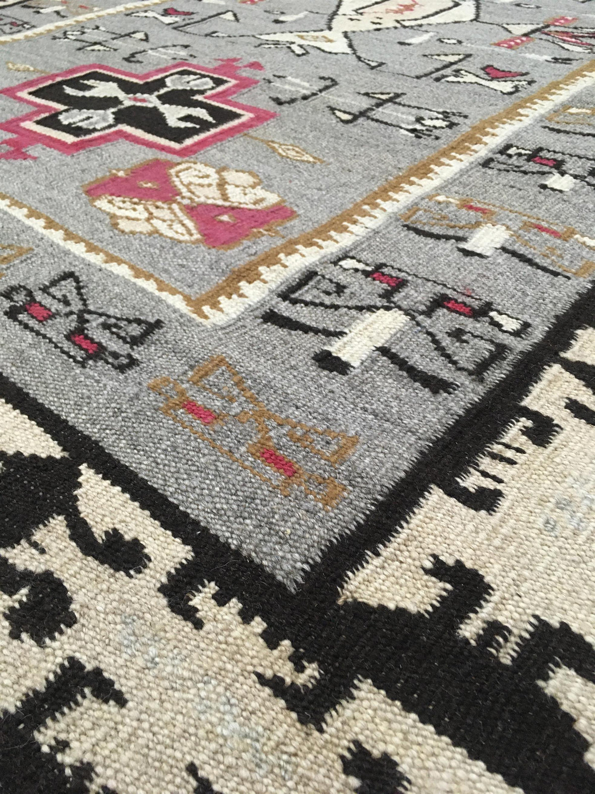 Close-up of a woven area rug showing a gray field with black, pink, beige, and white geometric motifs and borders. Atlantica Organics curates antique and handmade rugs, Persian, Afghan, Tibetan, Pakistani, Turkish Oushak, Kilim, Khotan, tribal and contemporary textiles for luxury interiors, homes and real estate. Our collections connect timeless craftsmanship with interior design, furniture, fine art, architecture and modern living. Explore curated rugs, collectible textiles, natural materials, cultural her