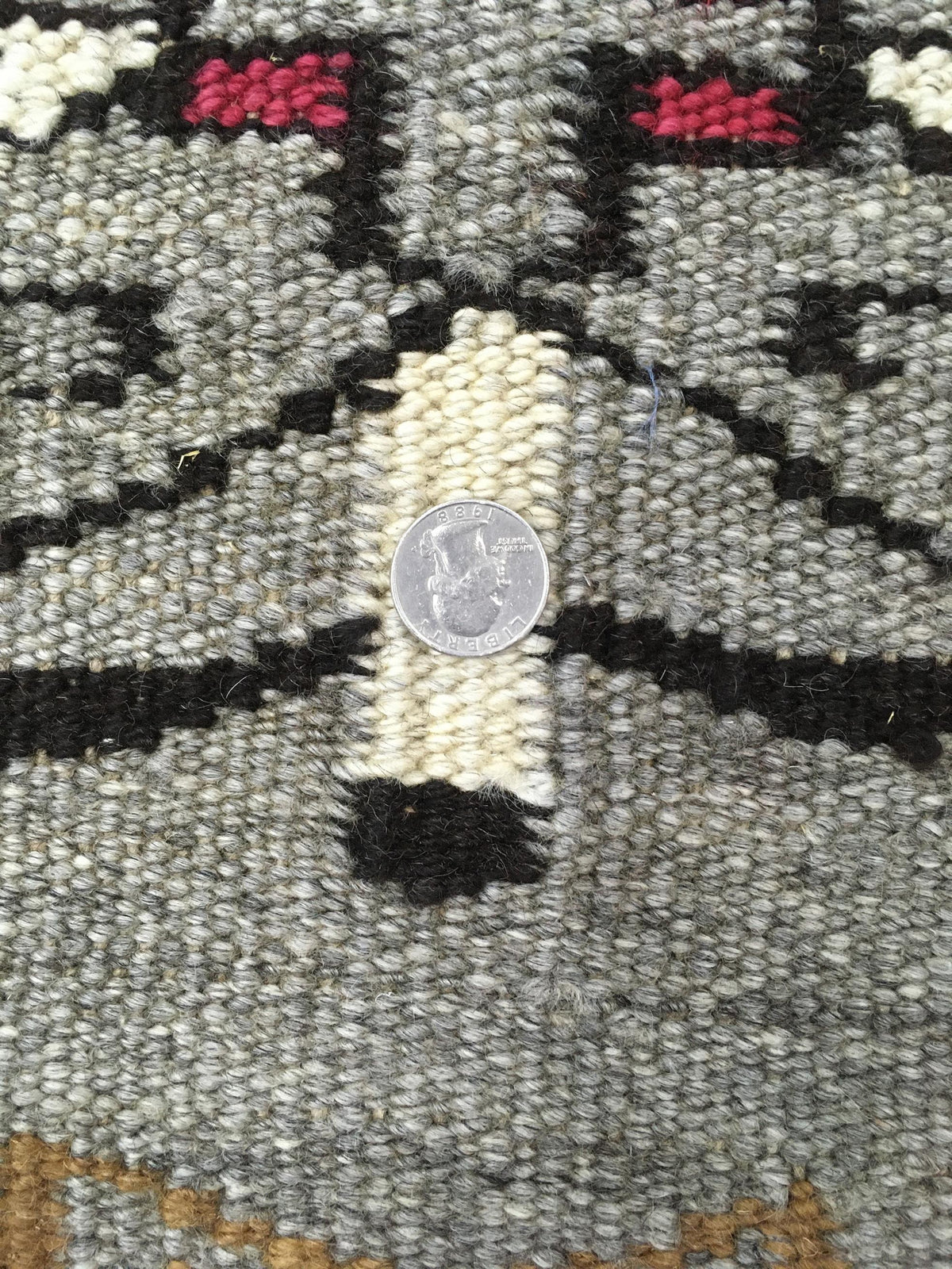 Close-up of a woven rug or textile with a gray base, black geometric lines, cream vertical bands, small red accents, and a coin placed in the center for scale. Atlantica Organics curates antique and handmade rugs, Persian, Afghan, Tibetan, Pakistani, Turkish Oushak, Kilim, Khotan, tribal and contemporary textiles for luxury interiors, homes and real estate. Our collections connect timeless craftsmanship with interior design, furniture, fine art, architecture and modern living. Explore curated rugs, collecti