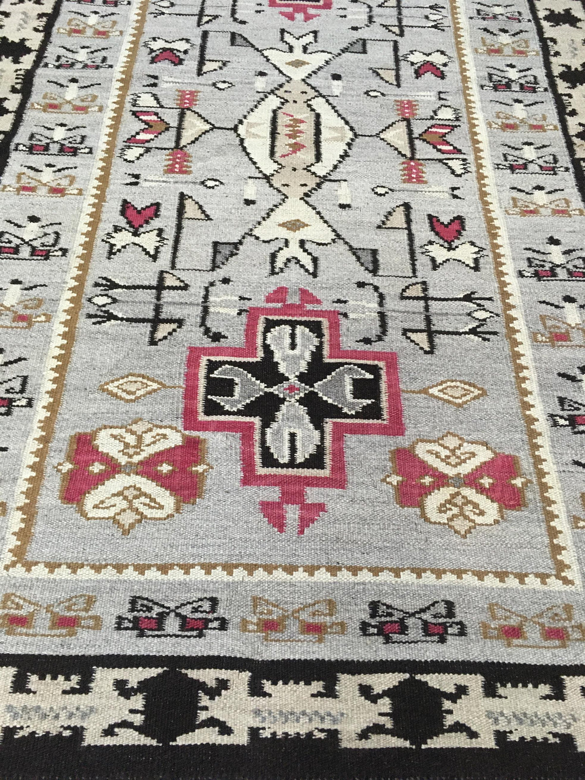 A close-up of a woven geometric rug with a light gray field, black, pink, beige, and white patterns, and a black border at the bottom. Atlantica Organics curates antique and handmade rugs, Persian, Afghan, Tibetan, Pakistani, Turkish Oushak, Kilim, Khotan, tribal and contemporary textiles for luxury interiors, homes and real estate. Our collections connect timeless craftsmanship with interior design, furniture, fine art, architecture and modern living. Explore curated rugs, collectible textiles, natural mat