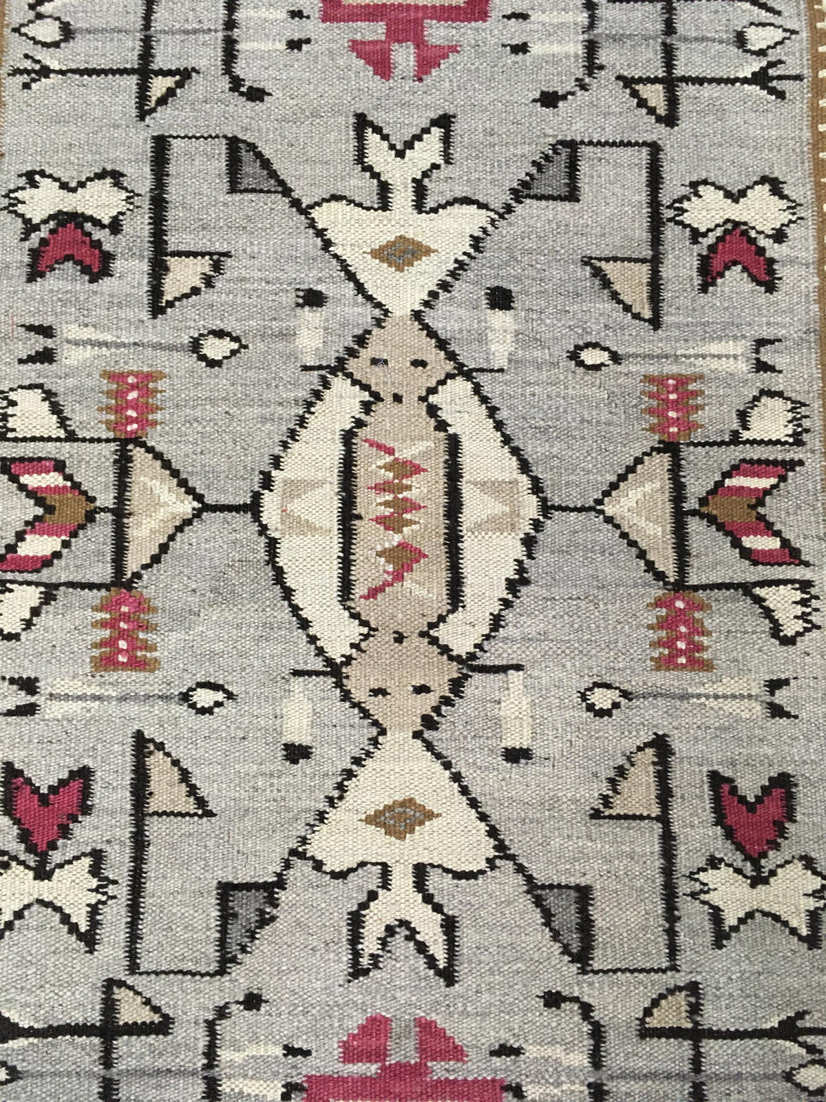 A close-up of a geometric patterned rug with a central diamond motif in cream, beige, and brown, surrounded by red, gray, and black tribal-style shapes on a light gray background. Atlantica Organics curates antique and handmade rugs, Persian, Afghan, Tibetan, Pakistani, Turkish Oushak, Kilim, Khotan, tribal and contemporary textiles for luxury interiors, homes and real estate. Our collections connect timeless craftsmanship with interior design, furniture, fine art, architecture and modern living. Explore cu