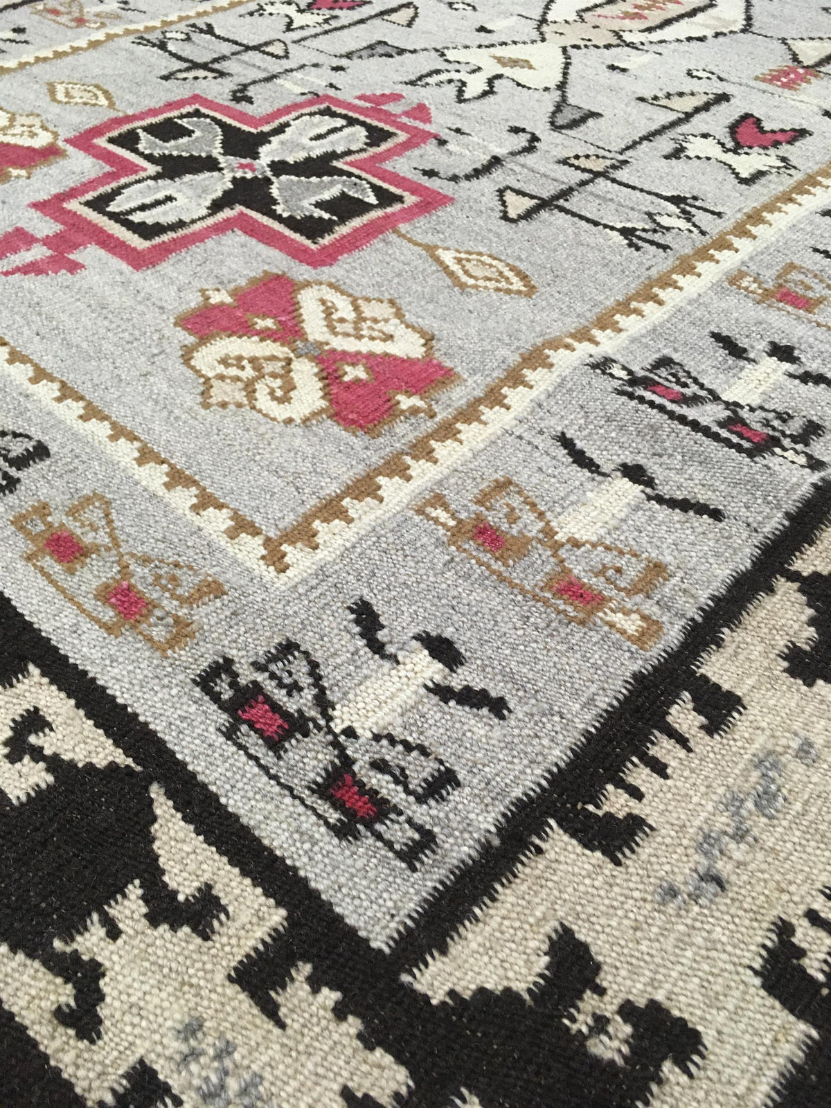 Close-up of a patterned area rug showing a light gray field with black, pink, beige, and white geometric motifs and borders. Atlantica Organics curates antique and handmade rugs, Persian, Afghan, Tibetan, Pakistani, Turkish Oushak, Kilim, Khotan, tribal and contemporary textiles for luxury interiors, homes and real estate. Our collections connect timeless craftsmanship with interior design, furniture, fine art, architecture and modern living. Explore curated rugs, collectible textiles, natural materials, cu