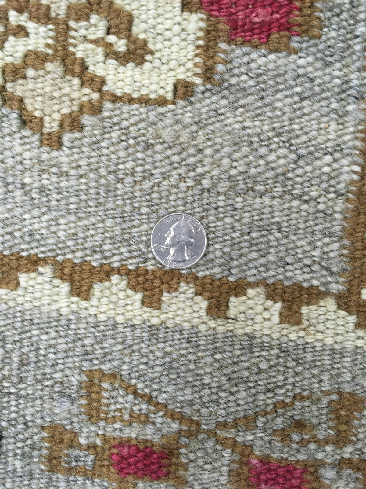 Close-up of a woven rug or textile with a geometric pattern in light gray, tan, cream, and pink, with a silver coin placed in the center for scale. Atlantica Organics curates antique and handmade rugs, Persian, Afghan, Tibetan, Pakistani, Turkish Oushak, Kilim, Khotan, tribal and contemporary textiles for luxury interiors, homes and real estate. Our collections connect timeless craftsmanship with interior design, furniture, fine art, architecture and modern living. Explore curated rugs, collectible textiles