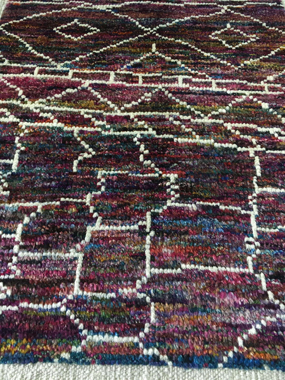 A close-up of a textured, multicolored woven rug with a geometric pattern in shades of purple, red, blue, green, and cream. Atlantica Organics curates antique and handmade rugs, Persian, Afghan, Tibetan, Pakistani, Turkish Oushak, Kilim, Khotan, tribal and contemporary textiles for luxury interiors, homes and real estate. Our collections connect timeless craftsmanship with interior design, furniture, fine art, architecture and modern living. Explore curated rugs, collectible textiles, natural materials, cul