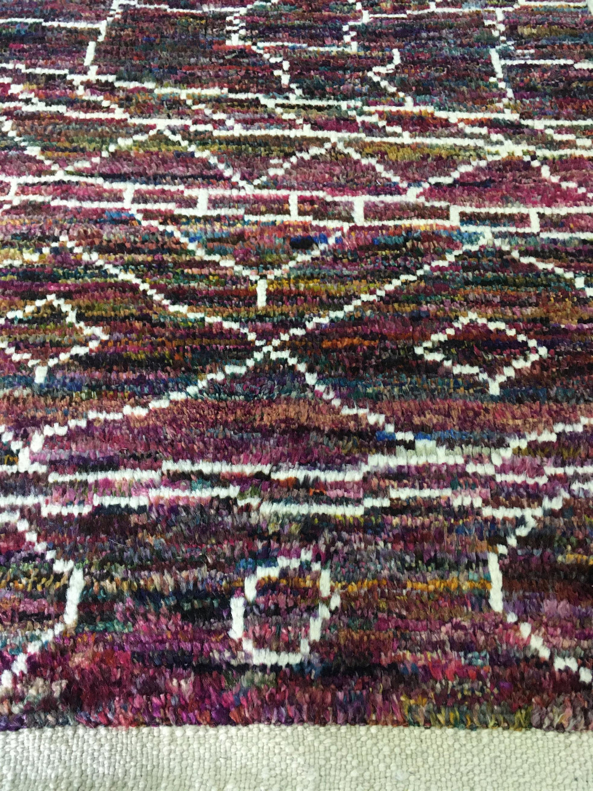 A close-up of a textured, multicolored woven rug featuring a geometric diamond pattern in shades of purple, red, blue, and yellow with white lines, finished with a cream-colored border at the bottom. Atlantica Organics curates antique and handmade rugs, Persian, Afghan, Tibetan, Pakistani, Turkish Oushak, Kilim, Khotan, tribal and contemporary textiles for luxury interiors, homes and real estate. Our collections connect timeless craftsmanship with interior design, furniture, fine art, architecture and moder