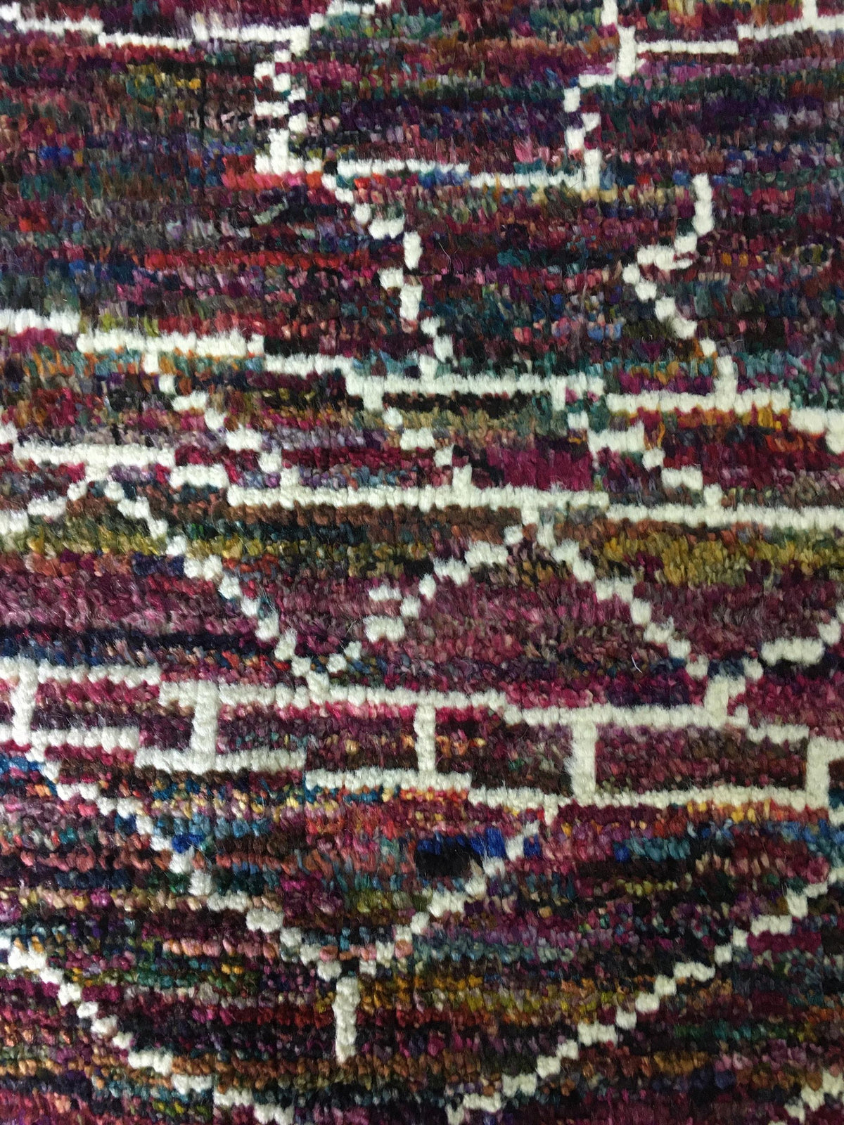 A close-up of a textured, multicolored rug featuring a geometric diamond pattern in shades of burgundy, purple, blue, green, and cream. Atlantica Organics curates antique and handmade rugs, Persian, Afghan, Tibetan, Pakistani, Turkish Oushak, Kilim, Khotan, tribal and contemporary textiles for luxury interiors, homes and real estate. Our collections connect timeless craftsmanship with interior design, furniture, fine art, architecture and modern living. Explore curated rugs, collectible textiles, natural ma
